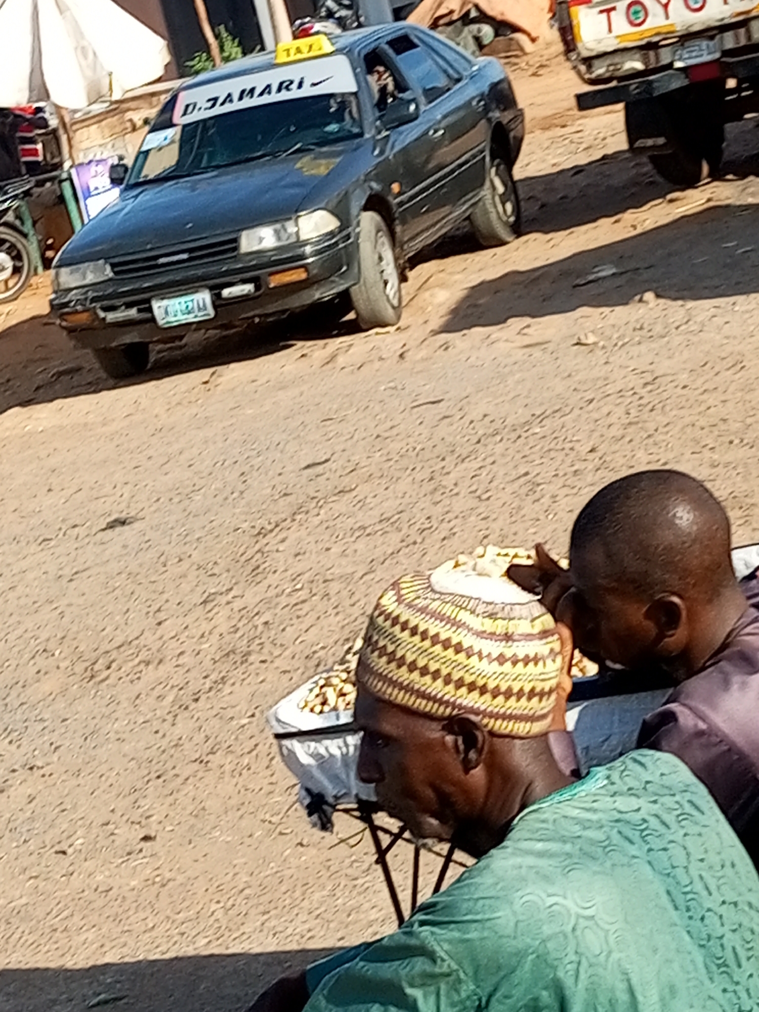 In Tumfure, Nigeria, a dark-colored taxi with "D.JAMARI" and a Nike logo on its windshield is parked on a dirt road. A yellow "TAXI" sign is visible on the roof. The taxi's license plate is partially obscured. In the foreground, two men are seated. One man, wearing a green patterned shirt and a yellow and brown cap, is looking down. The other man, wearing a dark purple shirt, is also looking down. Between them, a white plastic bag filled with what appears to be food items is placed on a small table or stand. In the background, another vehicle, possibly a Toyota truck, is partially visible. The ground is sandy and uneven. The scene appears to be outdoors, under natural light.