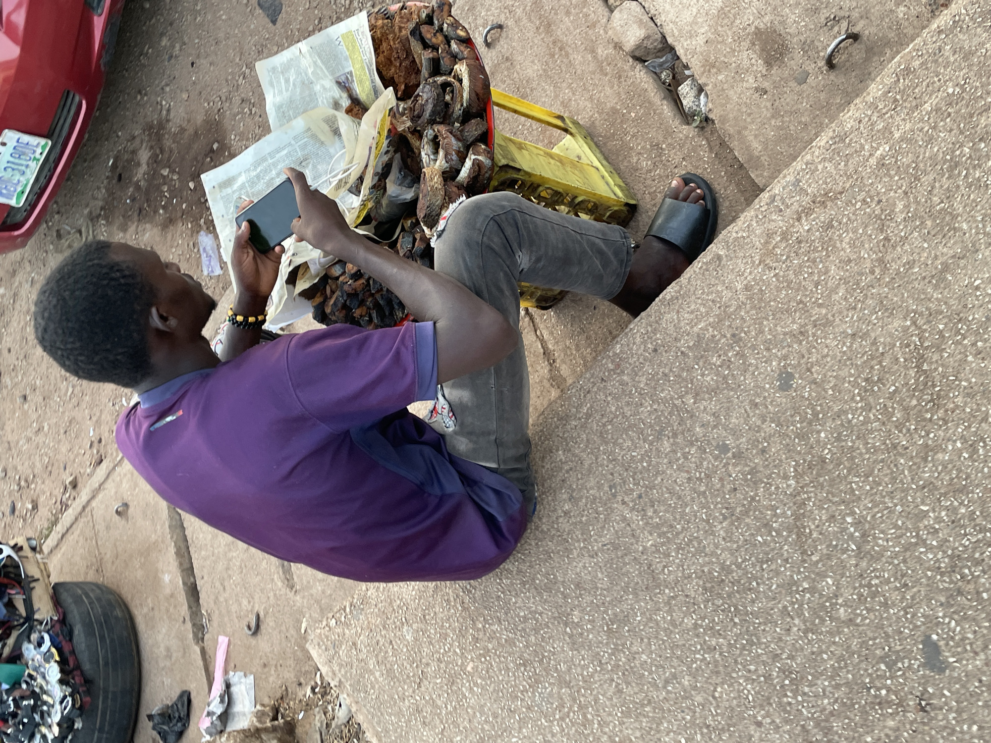 A man sits on a concrete ledge in Barakin Lerawi, Nigeria, looking at his phone. Next to him is a yellow crate containing dried fish wrapped in newspaper. A red car is parked nearby and a tire lies on the ground to the left.