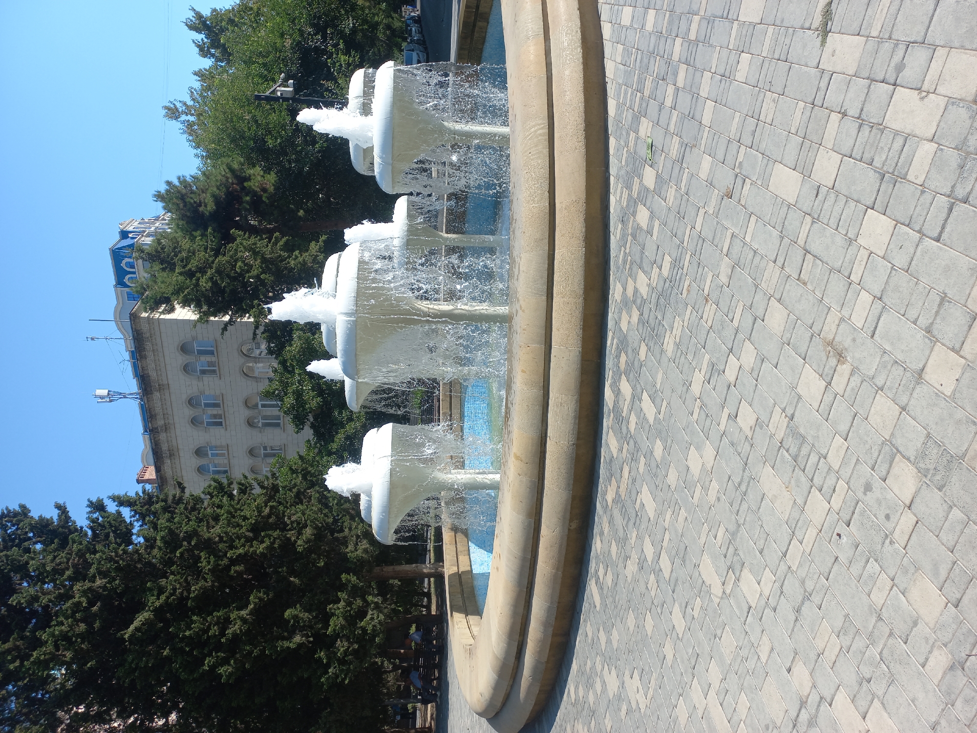The image depicts a fountain in a public space, likely a park or square, in Baku, Azerbaijan. The fountain has multiple tiered white structures from which water is actively spraying upwards, creating a dynamic visual effect. The fountain is set within a circular, beige stone basin. The ground surrounding the fountain is paved with interlocking grey and beige rectangular bricks, laid in a repeating pattern. In the background, lush green trees are visible, providing a natural contrast to the urban environment. Behind the trees, a building with an arched window facade and a blue-tiled roof is partially visible. The sky above is a clear, bright blue, indicating a sunny day. The scene is tranquil and showcases a typical urban amenity.