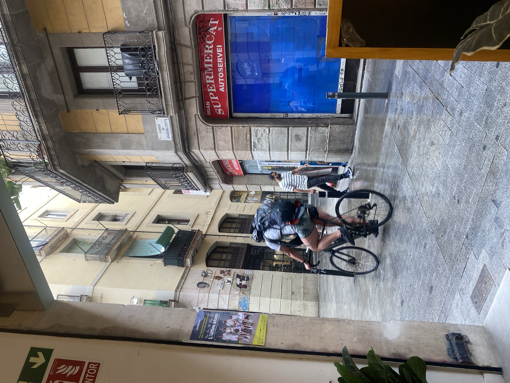 This image captures a vibrant street scene in an urban environment, likely Barcelona. The perspective is from a slightly elevated position, possibly from a building entrance or balcony, looking down a narrow street or alley.
The street is paved with intricate grey tiles, and lined with multi-story buildings featuring traditional architecture, numerous windows, and decorative iron balconies. Some balconies are adorned with green awnings.
In the mid-ground, a person wearing a backpack, shorts, and a light-colored shirt is actively cycling on a black bicycle, their motion creating a blur effect. Further down, another individual in a striped t-shirt and dark pants stands near a building entrance, partially obscured.
A prominent feature is a supermarket storefront with a red sign displaying "MIAN SUPERMERCAT AUTOSERVEI" (Mian Supermarket Self-service) above a striking bright blue window. Graffiti, including the text "RICKS MARE," is visible on the wall adjacent to the supermarket window. On a building to the left, a white street sign clearly reads "CARRER DE JOAQUÍN COSTA." Below the cyclist, a poster on a wall advertises "TEATREGOYA" and a show titled "L'últim àtom," with the tagline "Una tragicomèdia sobre el que som i de..." (A tragicomedy about what we are and...).
The scene is illuminated by daylight, suggesting it's either morning or afternoon. In the immediate foreground, parts of an interior space are visible, including a green plant and a partially cropped sign that includes "PROMOTOR" and a fire extinguisher symbol with "NTOR" (likely part of "EXTINTOR").