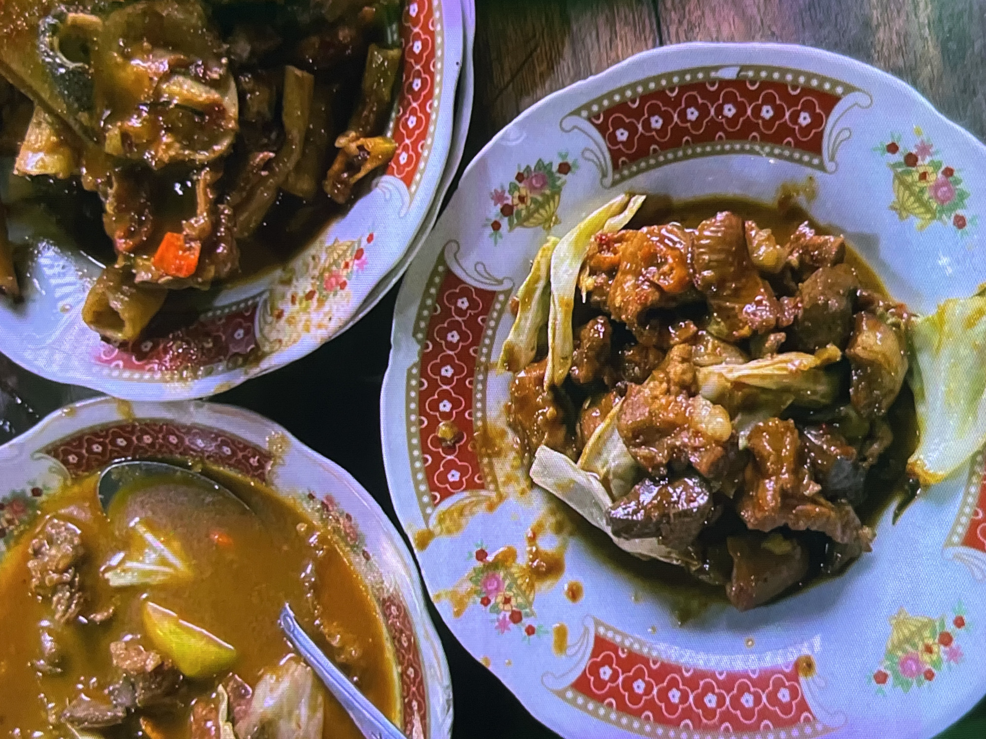 The image shows a top-down view of three plates of food on a wooden surface. The plates are white with a decorative red and white floral pattern around the rims and small floral accents scattered on the white surface. The food appears to be a savory stew or stir-fry.
The plate on the left contains a dish with chunks of meat and vegetables in a dark, glossy sauce. Some pieces of orange vegetable, possibly carrot, are visible.
The plate in the upper right is filled with pieces of meat in a similar dark sauce, mixed with some lighter-colored leafy vegetables, possibly cabbage.
The plate in the lower left is a bowl containing a rich, orange-colored broth with pieces of meat and what looks like slices of lime or lemon. A spoon is visible in the bowl.
The overall scene suggests a meal is being served or has just been eaten. The lighting is dim, and there are no people visible. The setting is likely a casual dining environment, possibly a restaurant or home. The dishes are typical of Indonesian cuisine, given the location context of Cadek, Indonesia. There is no visible text in the image.