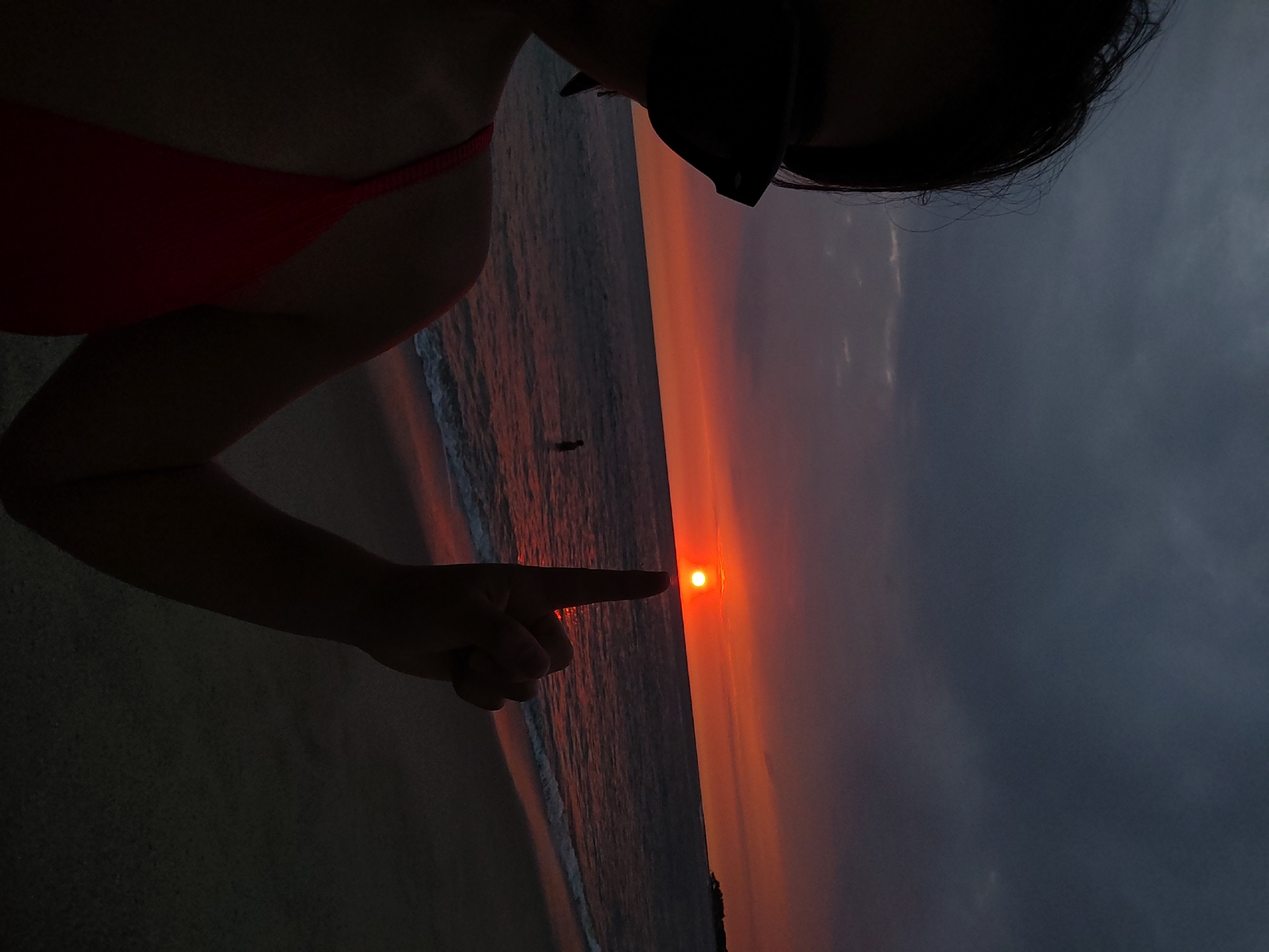 A person in a red swimsuit and sunglasses points a finger towards the setting sun. The setting sun is in the background along the coast of Bocapán, Peru. There is also a person swimming in the ocean.