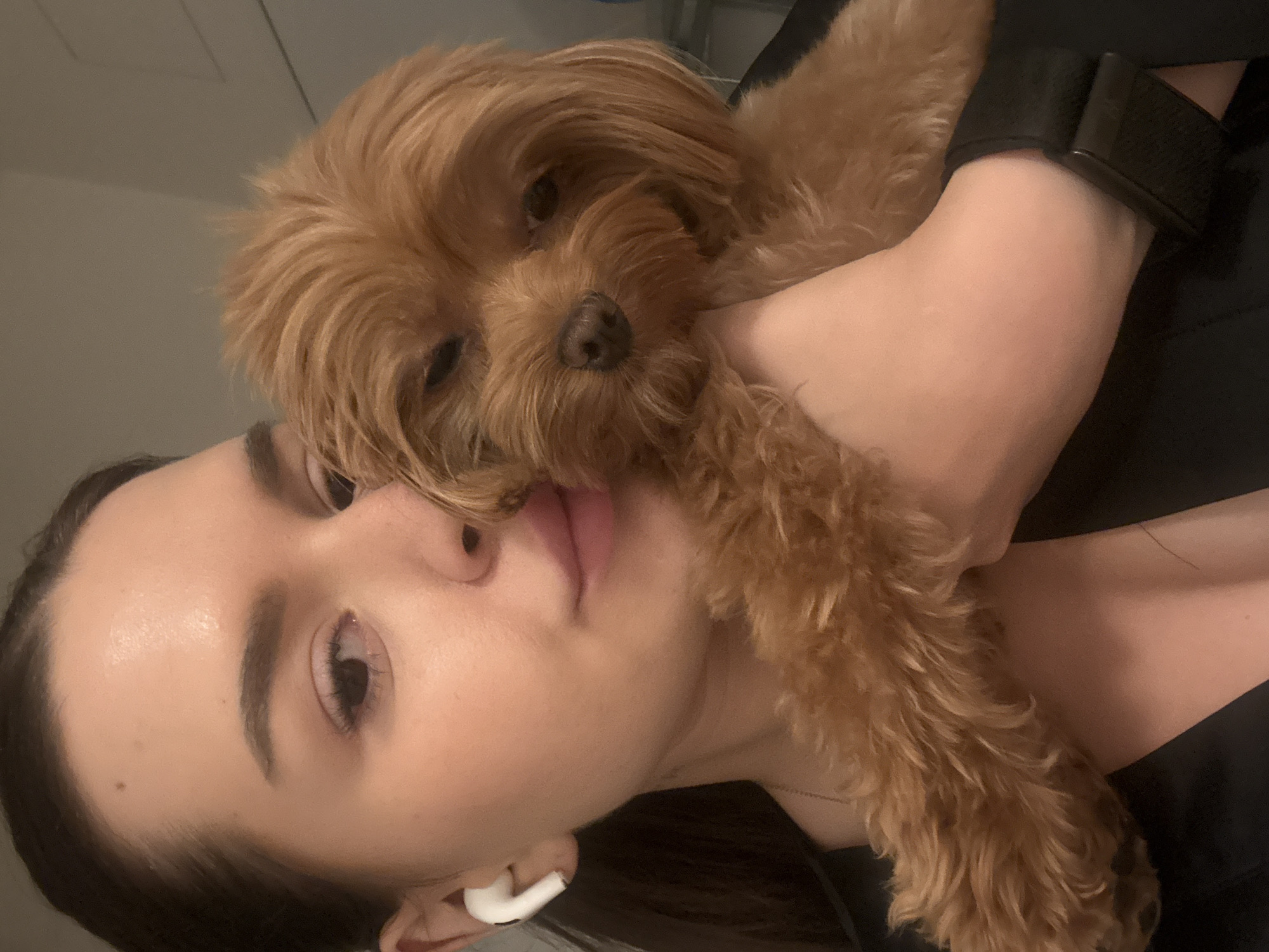 A woman with dark hair and fair skin is holding a small, fluffy, light brown dog. She is wearing a black top and a white wireless earbud in her left ear. The dog is nestled against her cheek and shoulder. The background is blurred and appears to be indoors. The scene is a close-up shot, focusing on the woman and the dog. There is no identifiable information that places the image in Prague, Czechia.