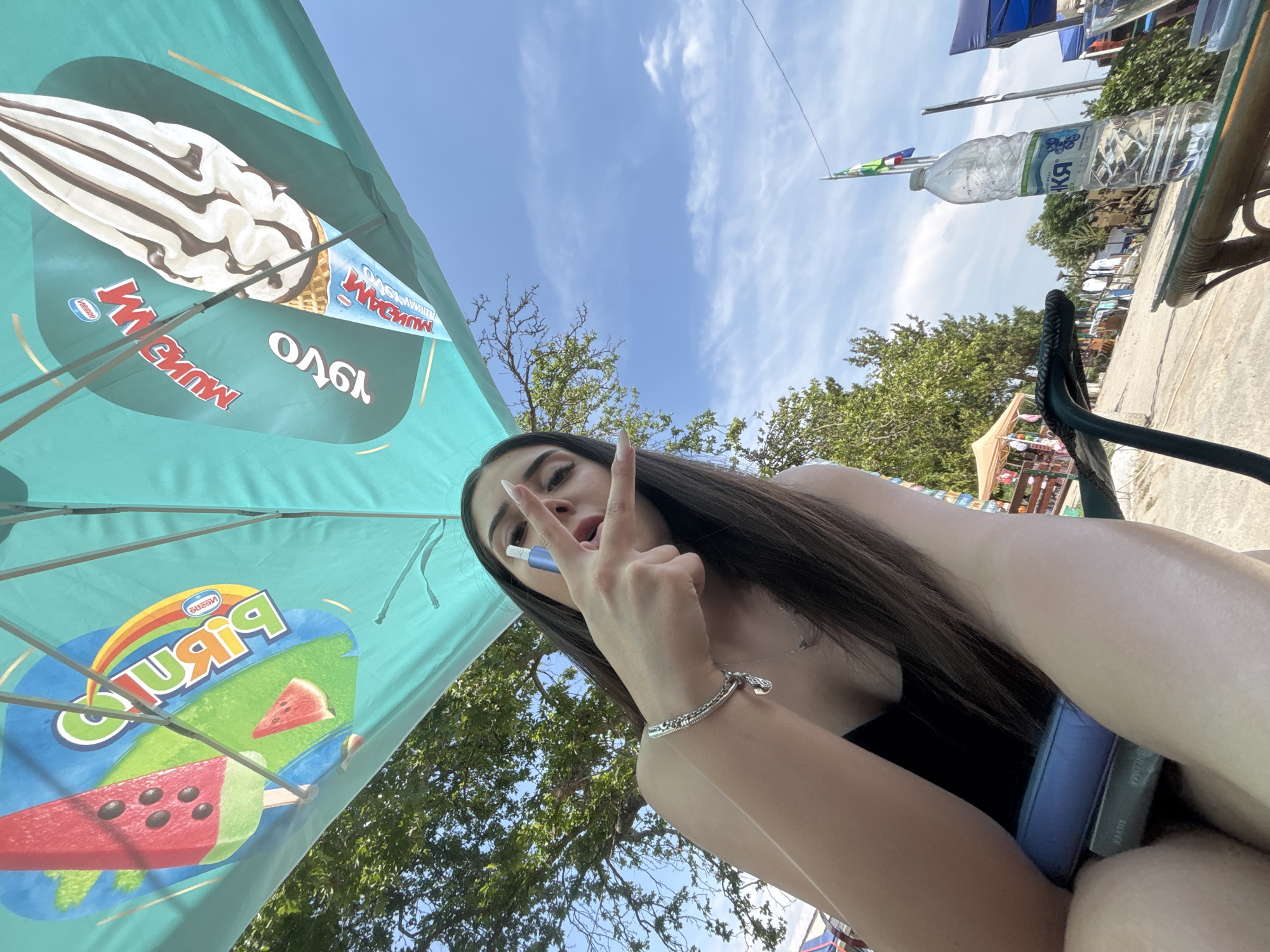 An outdoor, low-angle selfie depicts a young woman seated under a large, teal-colored promotional umbrella in Kostievo, Bulgaria. The woman, with long dark hair and wearing a black top, occupies the central-left foreground. Her left hand is raised, performing a peace sign gesture while holding a blue-and-silver cylindrical vape pen between her thumb and index finger. She has light-colored, elongated fingernails. A silver chain bracelet with a charm is visible on her left wrist, and a delicate silver necklace rests on her chest.
The umbrella, angled above the woman, displays two distinct upside-down ice cream advertisements. The left advertisement features a watermelon-shaped popsicle and a watermelon slice, branded "Биляна" (Bilyana) with a rainbow logo and "Nestle." The right advertisement shows a soft-serve ice cream cone with chocolate drizzle, branded "МЭГНУМ" (Magnum) and "Nestle."
The background reveals a clear blue sky with sparse white clouds. Lush green trees are prominent, suggesting a natural outdoor environment. To the far right, a transparent plastic water bottle with a partially visible "БУЛАРКА" label rests on a glass-top table with a woven base. Beyond the table, several market-like tents with varying canopy colors (light brown and blue) are discernible, situated along what appears to be a dirt or gravel path. A flagpole with a green and white flag is visible in the mid-ground, to the right.