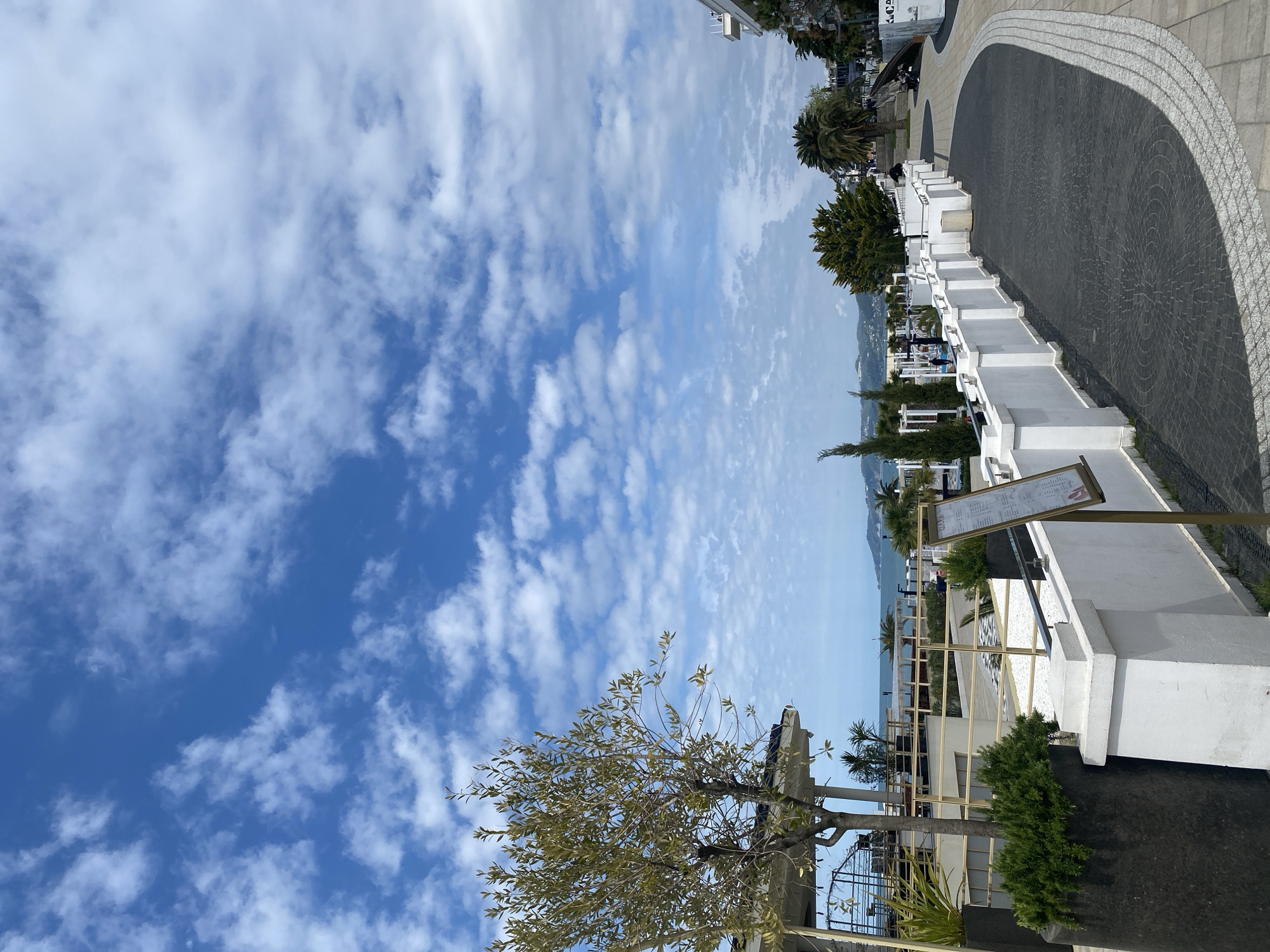 The image captures a bright, sunny daytime scene of a coastal promenade in what appears to be a resort town. The sky is a vivid blue with numerous scattered white clouds.
In the foreground and mid-ground, a meticulously designed outdoor area is visible. A dark grey paved walkway with a distinct semi-circular pattern curves through the scene, bordered by an elegant light grey and white patterned edge. Along this path, a series of low, white walls or planters are arranged, some containing small green bushes. Further back, various trees and plants punctuate the landscape, including several palm trees and another tree with lighter green foliage that dominates the lower left of the frame. Towards the right, a wooden sign on a stand is prominently displayed; it appears to be a menu or information board for an establishment, with text visible though illegible due to distance.
Beyond the immediate promenade structures, the calm, light blue expanse of the sea stretches out to the horizon. In the far distance, hazy mountains or hills can be seen, completing the scenic backdrop. A few indistinct figures are present in the mid-ground, suggesting people are strolling or relaxing in the area. The bright sunlight indicates clear weather, and the overall impression is one of leisure and beauty, consistent with a resort location like Sochi, Russia.