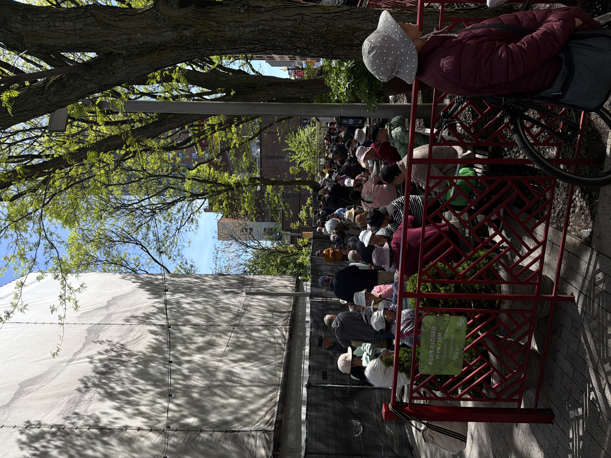 This image shows a group of people standing outside of a closed park. The park is bordered by a fence made of red metal with an Asian-inspired design. A sign is attached to the fence, indicating that the park is closed from 11 PM to 7 AM and that trespassers will be prosecuted. A large white sheet covers a section of the fence, and the shadows of tree branches cast patterns on the sheet. A dense crowd of people, mostly wearing hats or caps, is gathered behind the fence. Mature trees line the area, with their branches and leaves partially obscuring the view of the sky and nearby buildings. One person is positioned near the fence with a bicycle. The location is in Boston, United States of America.