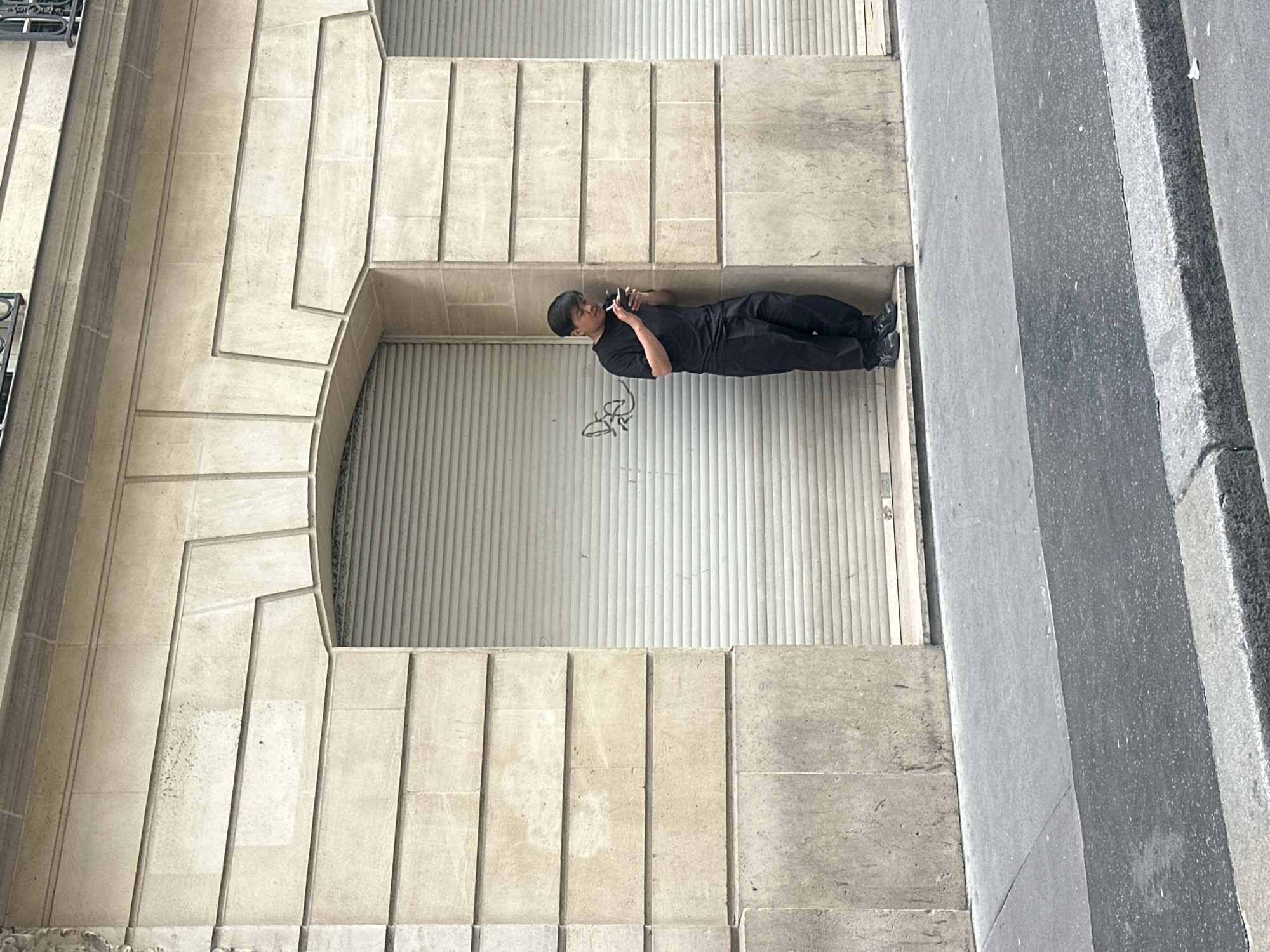 A medium-eye-level shot, taken from an angle, depicts a man in dark clothing standing in front of a shuttered storefront in Paris, France. The building's facade is constructed from light-colored stone blocks arranged in a regular pattern, with decorative cornices and moldings around the window and doorway. The man is holding a cigarette and a smartphone. The closed shutter is white with vertical ribbing and some graffiti. A sidewalk is visible along the right edge of the frame, leading towards the street.