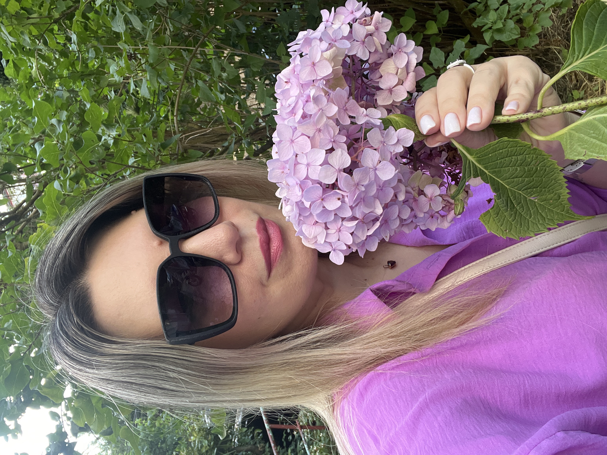 A medium shot captured in Sochi, Russia, features a woman holding a bouquet of light purple hydrangeas. She is wearing a vibrant pink collared shirt and large, dark sunglasses. Her blonde hair is partially visible, falling past her shoulders. The background consists of lush green foliage, suggesting an outdoor, natural setting. The woman's right hand, with neatly manicured nails, is extended forward, supporting the flowers. A light-colored strap from a bag is visible across her chest. The overall impression is a candid portrait taken outdoors.