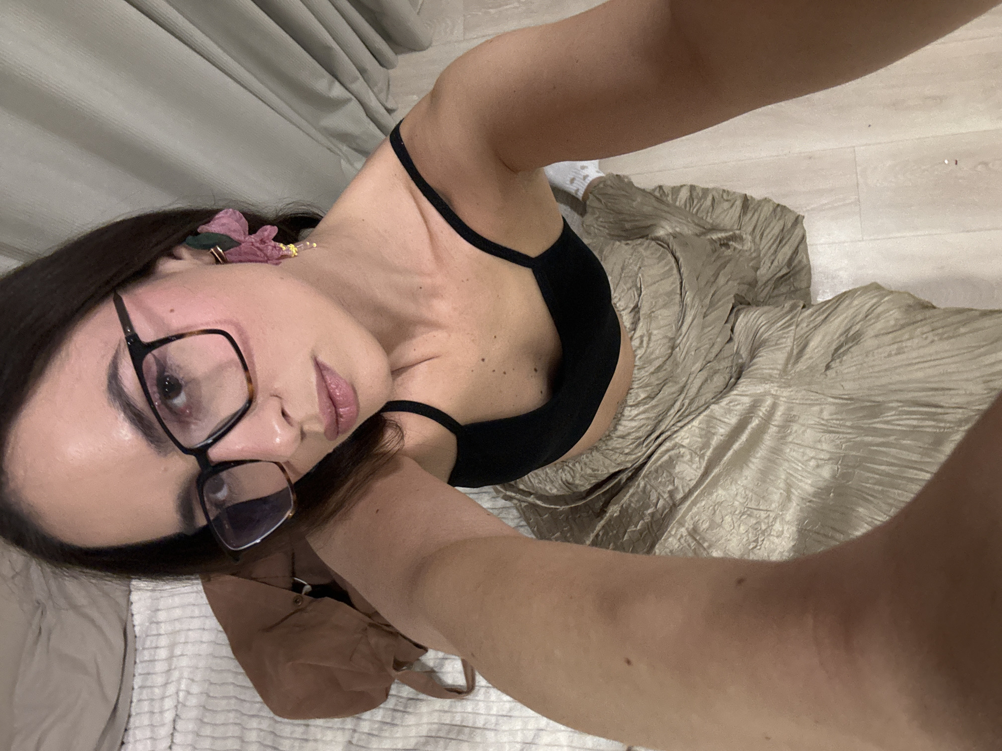 A person takes an upward-facing selfie in an indoor setting, captured from a high angle looking down. The individual, appearing to be an adult female with long dark hair, wears black framed glasses and a black bralette or crop top. A light gold or champagne-colored pleated skirt is visible below her waist. A decorative hair clip or earring, featuring a purple/pink flower with a gold accent, is visible near her ear. Her lips have a subtle pink color, and she looks directly towards the camera with a neutral expression.
The background features light-colored wooden flooring and textured light gray curtains. A white, textured blanket or rug is partially visible in the lower left corner, along with a brown bag or piece of clothing. A white sock can be faintly seen on the floor near her leg. No text is visible in the image. The lighting appears to be artificial. The setting is an indoor space, likely a room within a residence. The location is Krasnoyarsk, Russia.