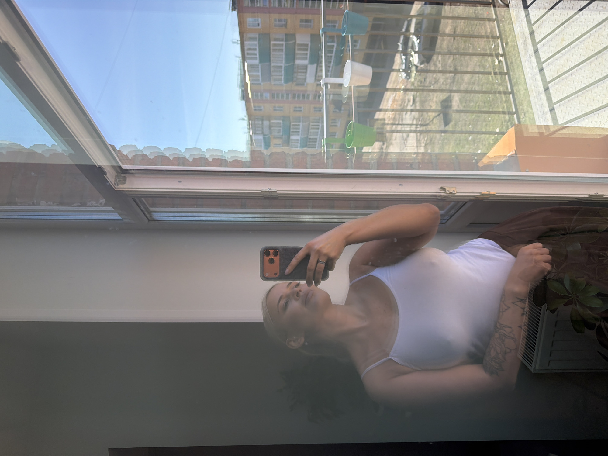 This image captures a vertical selfie reflection of a woman in a window or glass door, rotated 90 degrees counter-clockwise. The woman, with blonde hair, is looking towards the camera and holding a smartphone with a dark case (featuring orange/brown buttons) to take the photograph. She is wearing a white-strapped top and has a visible tattoo on her left arm.
The reflection divides the view into foreground (the woman) and background (the outdoor scene). Behind the woman's reflection, the indoor setting appears dark, possibly a floor or dark wall. Above her head in the reflection, a white ceiling or wall is visible.
Through the window, a clear blue sky is visible at the top. The mid-ground reveals a multi-story apartment building with numerous windows and balconies, featuring a cream/yellow facade. Hanging outside the window are three small, colorful planters or cups in light blue, white, and green. Below the building, an outdoor area with a metal fence or railing casts distinct shadows on the ground, indicating bright daylight. A cardboard box rests on the exterior windowsill. To the woman's right in the reflection, an indoor plant and a white air conditioner unit or vent are visible. The overall scene suggests a residential setting in a sunny daytime environment, consistent with urban areas like Sarapul, Russia. There is no discernible text in the image.