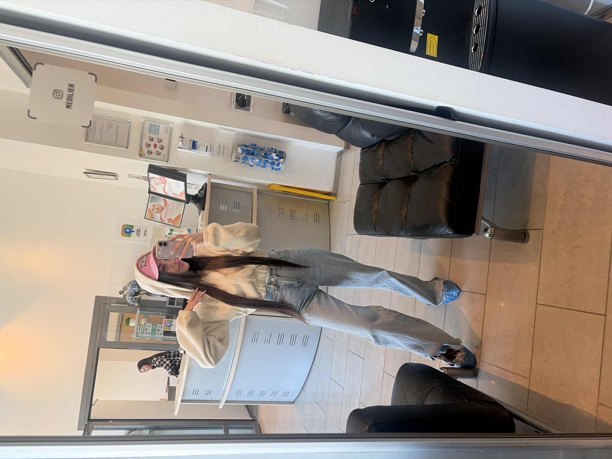 A young woman takes a mirror selfie indoors, likely in a waiting area or reception of a clinic or aesthetic center in Saint Petersburg, Russia. She is reflected full-body in a large mirror, standing with her smartphone held up to capture the image. She wears a light-colored, possibly cream, hooded fleece jacket, light-wash blue jeans, and a light pink baseball cap worn backward. Her long, dark hair falls over her shoulders. Over her shoes, she has put on blue disposable shoe covers, indicating a requirement for hygiene within the facility.
The setting visible in the reflection includes a minimalist white reception desk or counter. On a wall behind the desk, a white sign with a black Instagram logo prominently displays "MEDILIER." Below it are several framed documents or certificates, along with various brochures and dispensers. A clear bag containing more blue shoe covers is visible on the counter. To the right, there are dark-colored, possibly leather, upholstered chairs or sofas, typical of a waiting room. Further back, a large black appliance, possibly a water cooler or coffee machine, has a yellow label with "HOT" partially visible. The floor is covered with light-colored square tiles. In the background reflection, another person's partial reflection is seen at the counter. The woman in the selfie has a relaxed expression, with a slight smile. The overall lighting suggests an artificial indoor environment.