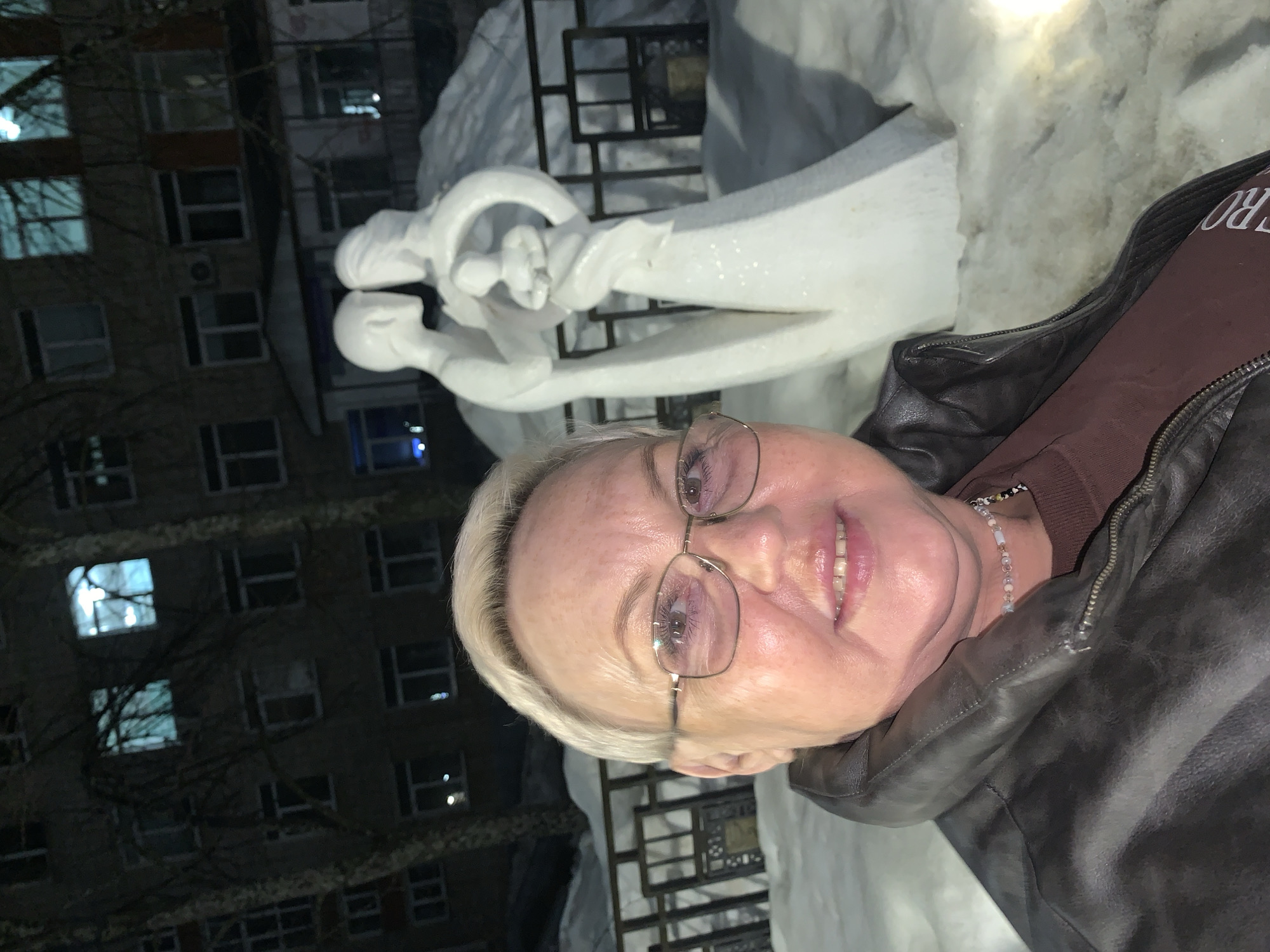 A smiling, middle-aged woman with short, light blonde hair and glasses is captured in a nighttime selfie. She wears a dark brown leather jacket over a reddish-brown top and a light-colored beaded necklace. Behind her, a white, stylized snow sculpture depicts a family: a male and female figure embracing a small child figure between them. The ground is covered in snow, with a dark metal fence visible behind the sculpture. In the background stands a dark, multi-story residential building with several windows illuminated, and bare tree branches are discernible against its facade. This scene is situated outdoors in Chaikovsky, Russia, during a winter period.