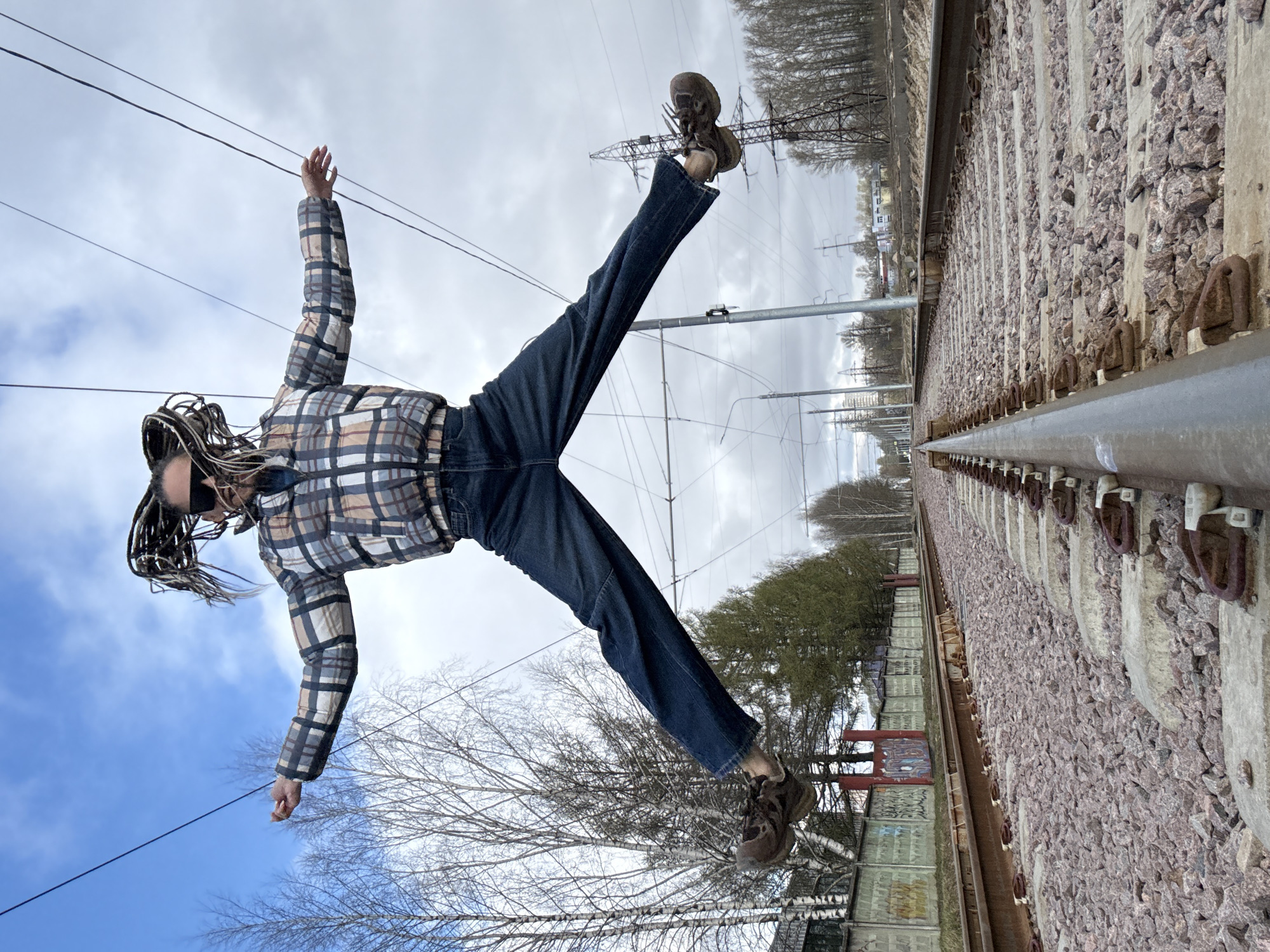 The image depicts a person suspended in the air between train tracks on one side and a blue sky with bare trees on the other. The person is wearing a plaid jacket, jeans, and brown boots. Their arms are outstretched, and their legs are spread apart in a V shape. The location is identified as Rusanovo, Russia. The train tracks appear to be actively used, with ballast and metal rails visible. Overhead electrical lines are present.