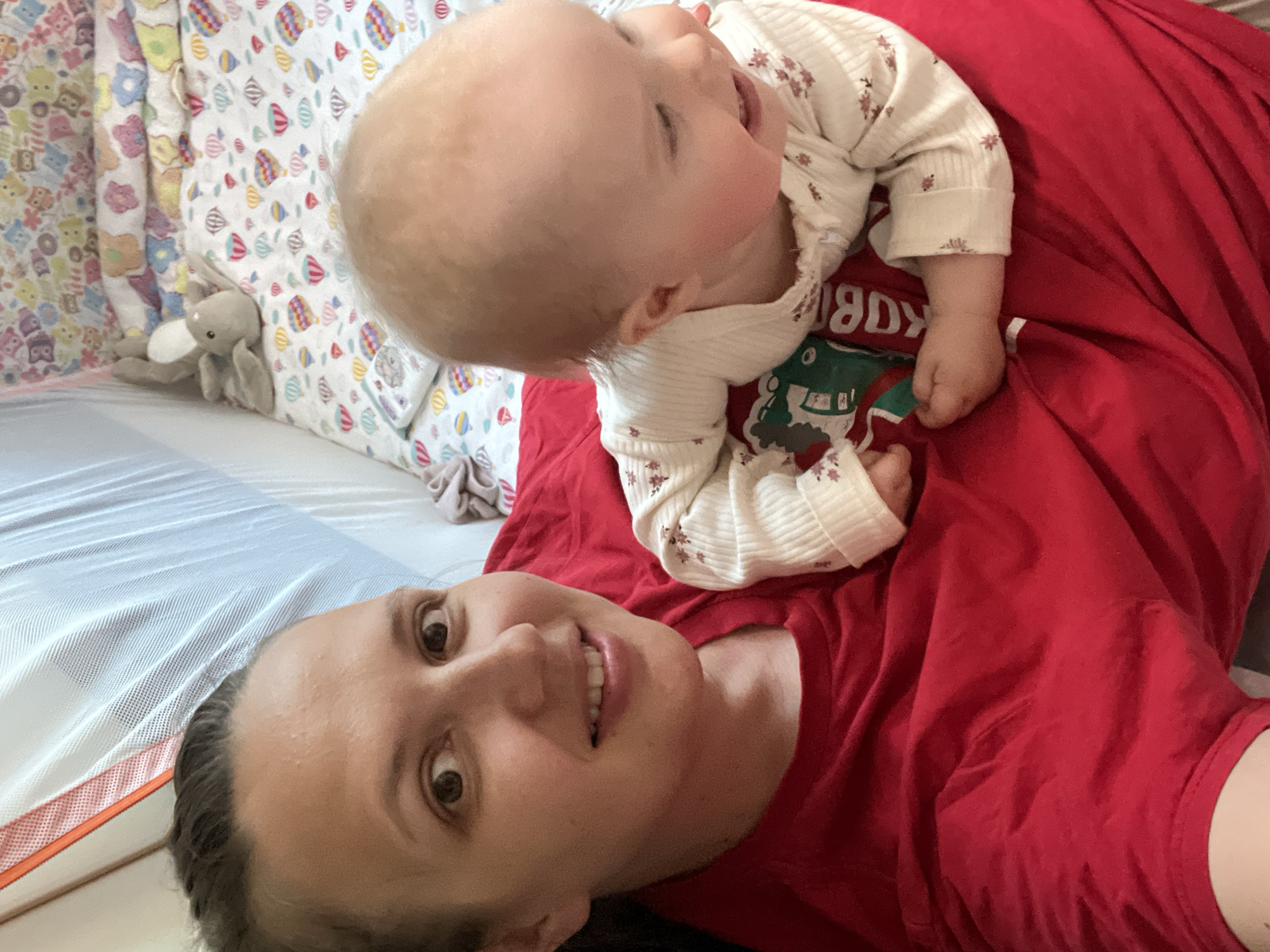 In an indoor setting, likely a child's crib or playpen, a woman and a baby are pictured. The woman is on the left side of the frame, her face close to the camera, and she is wearing a red t-shirt. Her expression is a mix of a smile and an open-mouthed gaze. Beside her, to the right, is a baby, also facing towards the right. The baby is wearing a light-colored onesie with a floral pattern and appears to be smiling and looking away from the camera. The background includes a white mesh netting on the left and a colorful bedding with hot air balloon and animal motifs on the right. A stuffed bunny toy is visible on the bedding. The overall scene suggests a moment of playful interaction between the woman and the baby. The location is identified as Maksimovo, Russia.