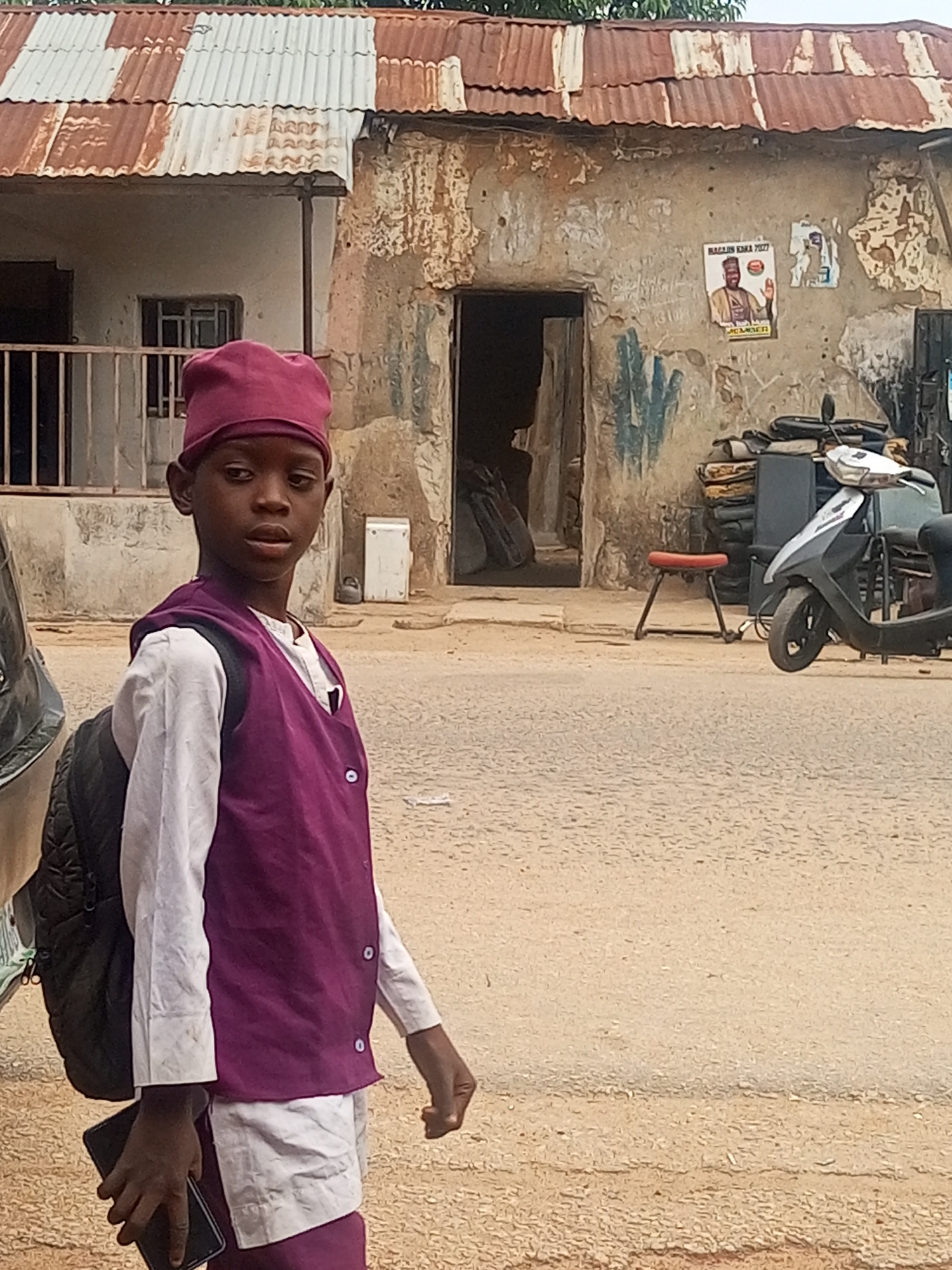A young boy wearing a purple cap, vest, and white shirt stands on a dirt road in Maiburiji, Nigeria. He is carrying a black backpack and holding a black smartphone. Behind him are buildings with corrugated metal roofs and weathered walls. Posters are affixed to one building. A scooter is parked nearby. The environment suggests a modest residential or commercial area in the city.