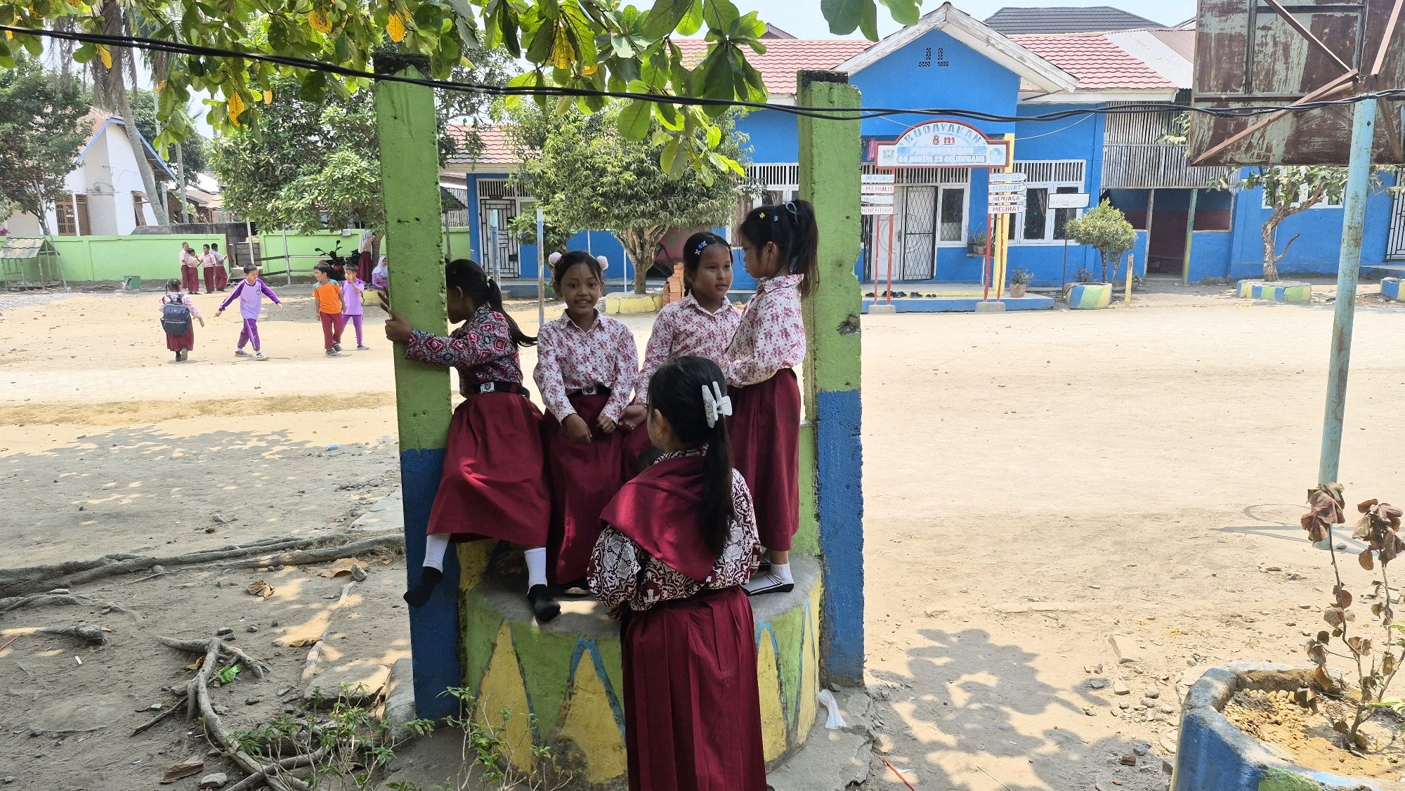 The image depicts an outdoor scene in front of a blue school building with a red roof in Putak, Indonesia. Several young girls, dressed in maroon skirts and patterned blouses, are gathered around a concrete pillar painted with yellow, green, and blue sections. Five girls are visible in the foreground and mid-ground. One girl is leaning against the pillar, another is partially visible to the left, and three are standing together on top of the pillar. A sixth girl, wearing a maroon cape and skirt, stands facing away from the camera. In the background, other children in school uniforms are walking or standing on the dusty schoolyard. The environment includes trees, a fence, and the school building. The overall impression is a group of students interacting in a school setting.