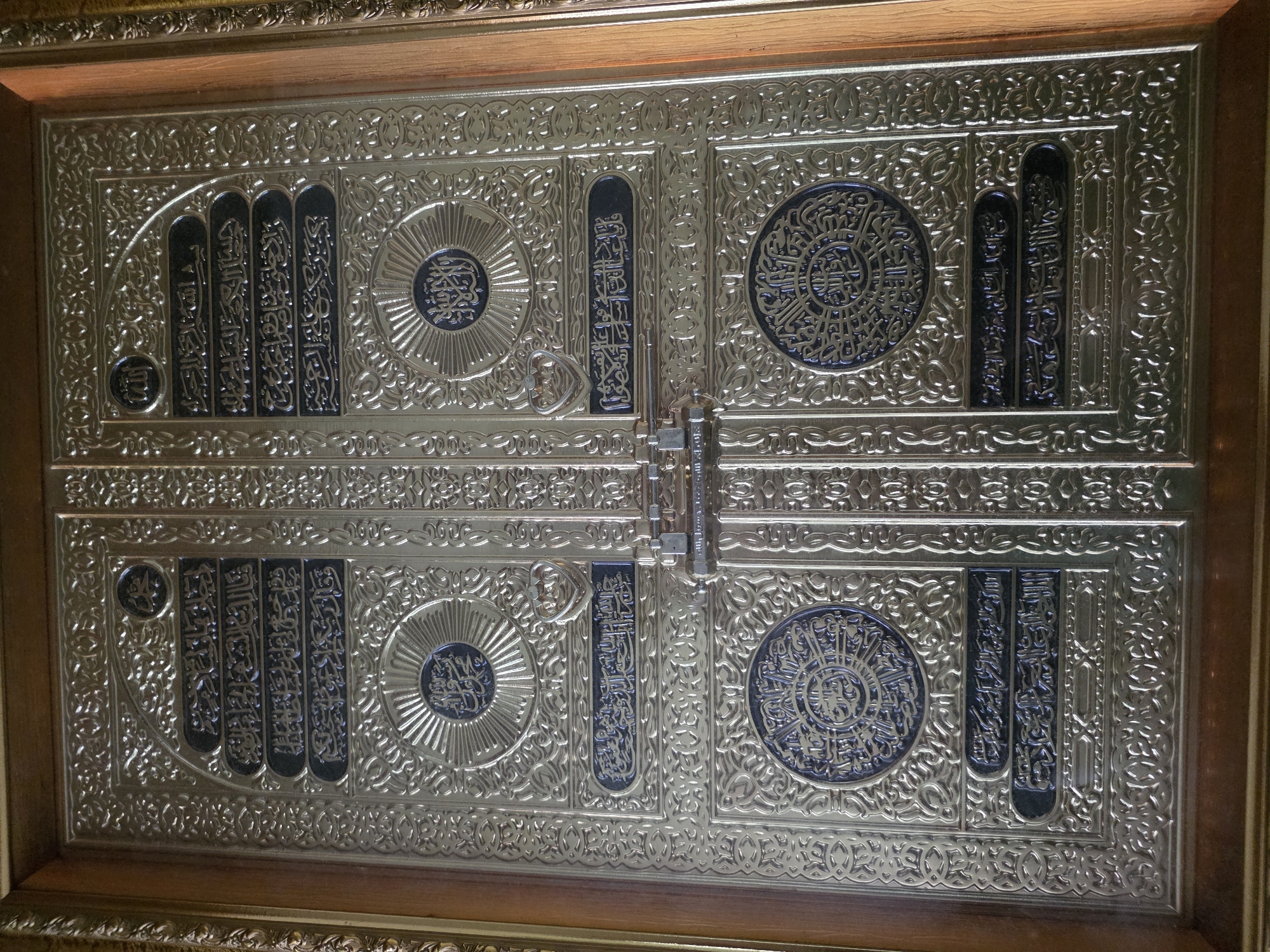 The image shows a close-up of an ornate, golden door with intricate carvings and Arabic calligraphy. The door appears to be a replica or a decorated facade, possibly part of an Islamic cultural display. The golden surface is highly reflective, highlighting the detailed patterns and inscriptions. There are circular emblems with radiating lines and central calligraphy, interspersed with horizontal bands of text. A metal latch mechanism is visible on the right side of the door. The door is set within a wooden frame. The location of Bireuenmeunasahblang, Indonesia, is not evident from the image itself, as the image only depicts the door and its immediate surroundings.