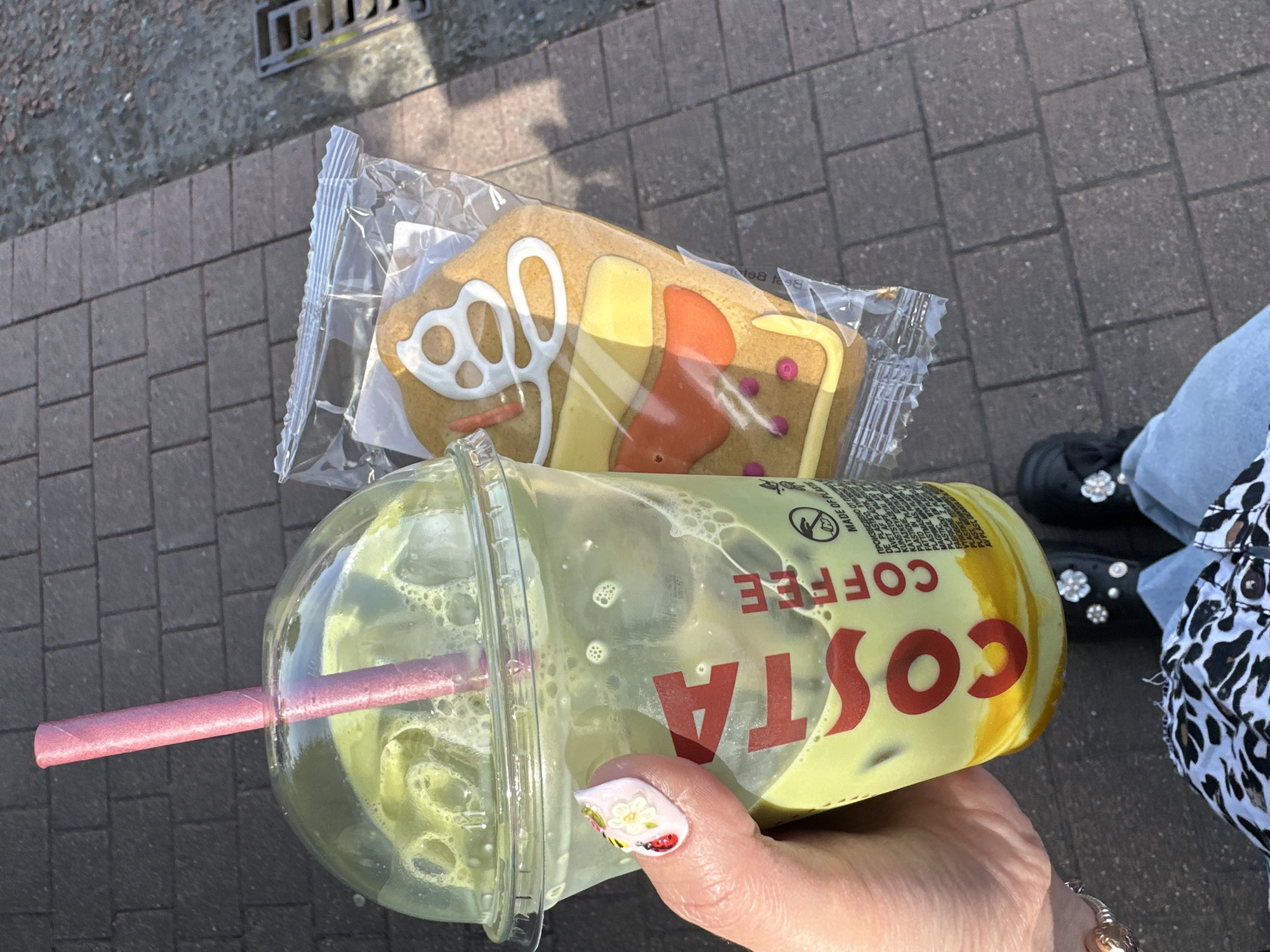 A hand holds a clear plastic cup containing a green beverage with ice and bubbles, topped with a domed lid and a pink straw. The cup has red lettering that reads "COSTA COFFEE." Next to the cup, also held by the hand, is a gingerbread cookie in a clear plastic wrapper. The cookie is decorated to resemble a house with white icing, yellow icing for a window frame, and orange icing with pink dots. The background is a paved surface made of dark grey rectangular bricks. The scene is outdoors, with sunlight casting shadows. The person is wearing blue jeans and black shoes with decorative floral embellishments. A black and white leopard print fabric is visible at the bottom of the image.
