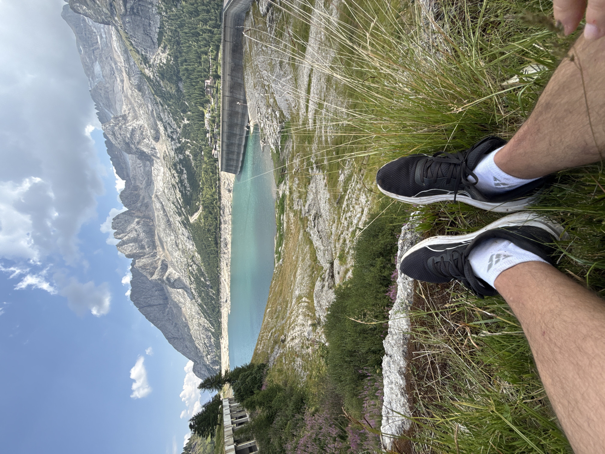 The image captures a scenic view of the Fedaia dam and lake in the municipality of Canazei, Italy. In the foreground, a person's legs, clad in black running shoes and white socks, are positioned on a grassy embankment. The background features the expansive turquoise waters of Lago di Fedaia, bordered by a large concrete dam. Beyond the dam, a lush green forest gives way to the rugged, rocky peaks of the Marmolada mountain range, with some snow visible on the highest summits. The sky is a clear blue, dotted with white clouds. The overall scene depicts a tranquil natural landscape with human presence indicated by the footwear.
