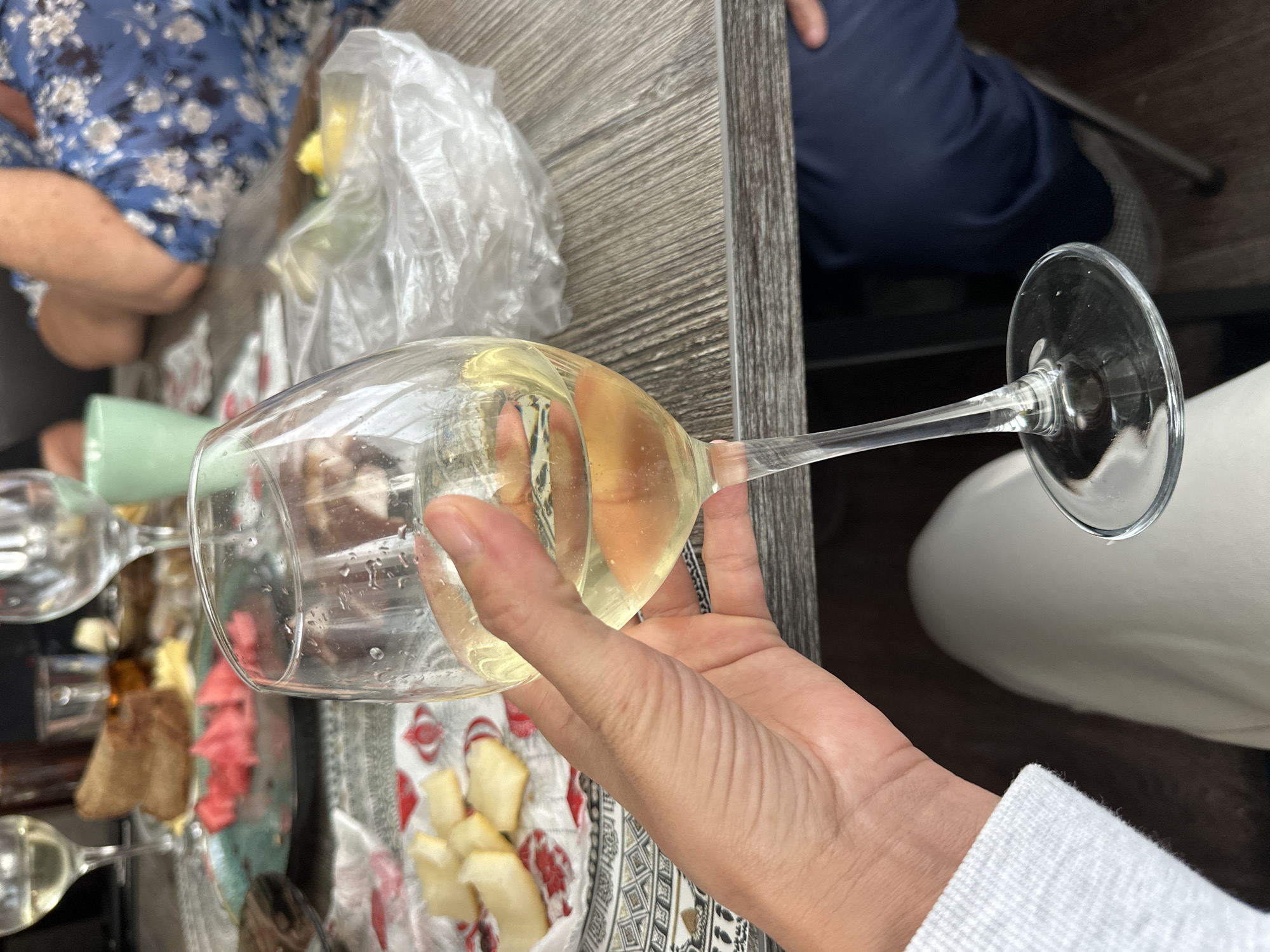 A first-person perspective shot shows a hand holding a wine glass filled with white wine. The glass is tilted slightly, with a few bubbles visible in the liquid. In the background, a table is laden with food and drinks, including a plate of watermelon slices, some bread, and small pieces of cheese. A plastic bag is also visible on the table. The setting appears to be an outdoor gathering in Baklashi, Russia, with a woman in a blue floral dress visible on the right side of the frame. The table surface is made of wood. The overall atmosphere suggests a casual meal or picnic.