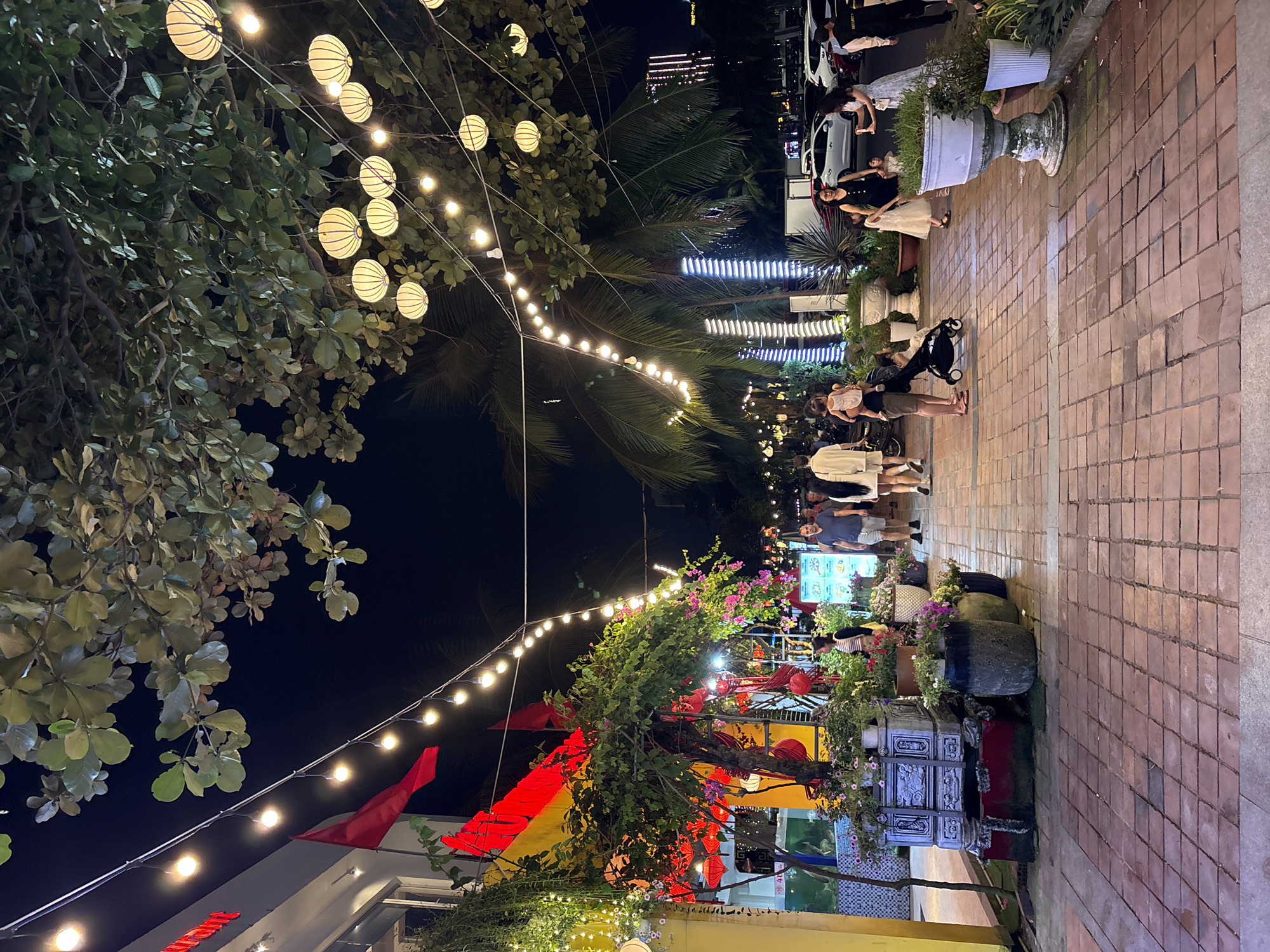 The image captures a vibrant, illuminated outdoor pedestrian area at night, likely a promenade or a decorated street in Da Nang, Vietnam. Numerous people, including adults and children, are seen strolling leisurely along a brick-paved pathway that extends from the foreground into the background on the right. One person is pushing a baby stroller.
The scene is richly decorated with various types of lighting and greenery. Overhead, string lights with bare bulbs stretch across the pathway, interspersed with larger, spherical white lanterns that hang from trees on the left. These trees feature dense, green foliage, and palm trees are also visible in the mid-ground. Red flags or banners are strung along the path, adding splashes of color. In the middle distance, vibrant red traditional lanterns are part of the decor, alongside lush potted plants and flower beds featuring pink and purple blooms. Large, ornate planters, some resembling classical urns, line the pathway.
In the background on the right, modern vertical strip lights illuminate structures, contrasting with the traditional lanterns. A white car with "SECURITY" visible on its side is parked further up the path. Another bright display board, possibly a menu for a nearby establishment, is visible in the mid-ground. The overall atmosphere is festive and lively, suggesting an enjoyable evening event or a popular gathering spot. The dark night sky highlights the warm glow of the lights, enhancing the inviting ambiance.