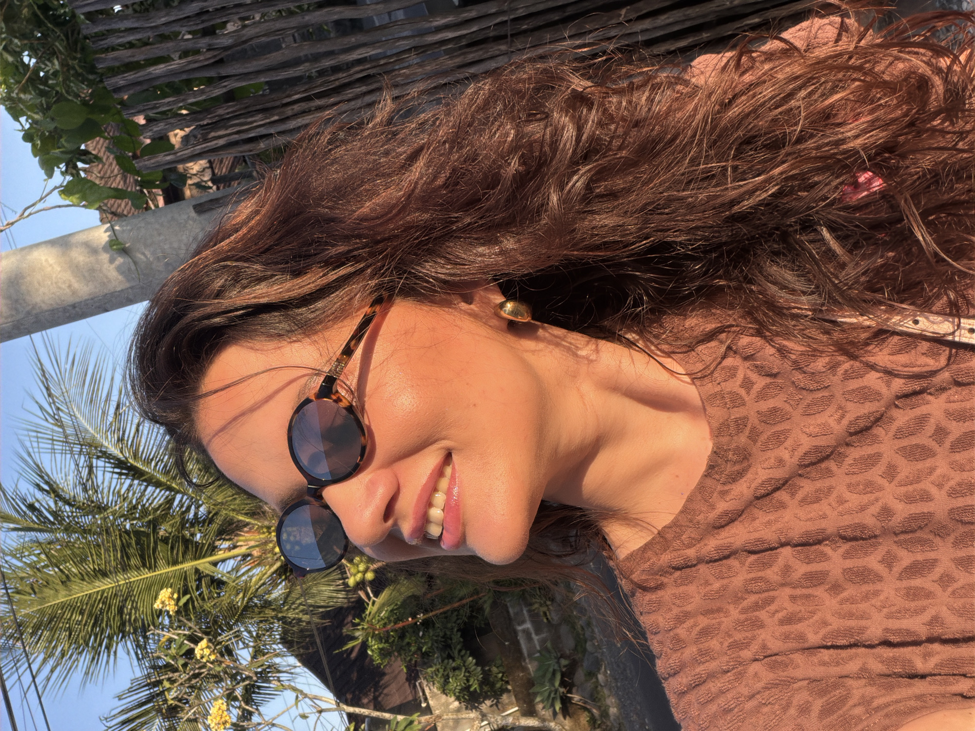 A woman with long, wavy brown hair and sunglasses smiles broadly while looking up and to her left, illuminated by golden hour sunlight. She is wearing a textured, rust-colored top and a gold earring. The background shows a rustic wooden fence, lush green foliage, and a palm tree, suggesting a tropical or subtropical setting. A utility pole is visible to the right of her face. This scene likely takes place in a residential area of North Kuta, Indonesia, given the vegetation and construction style.