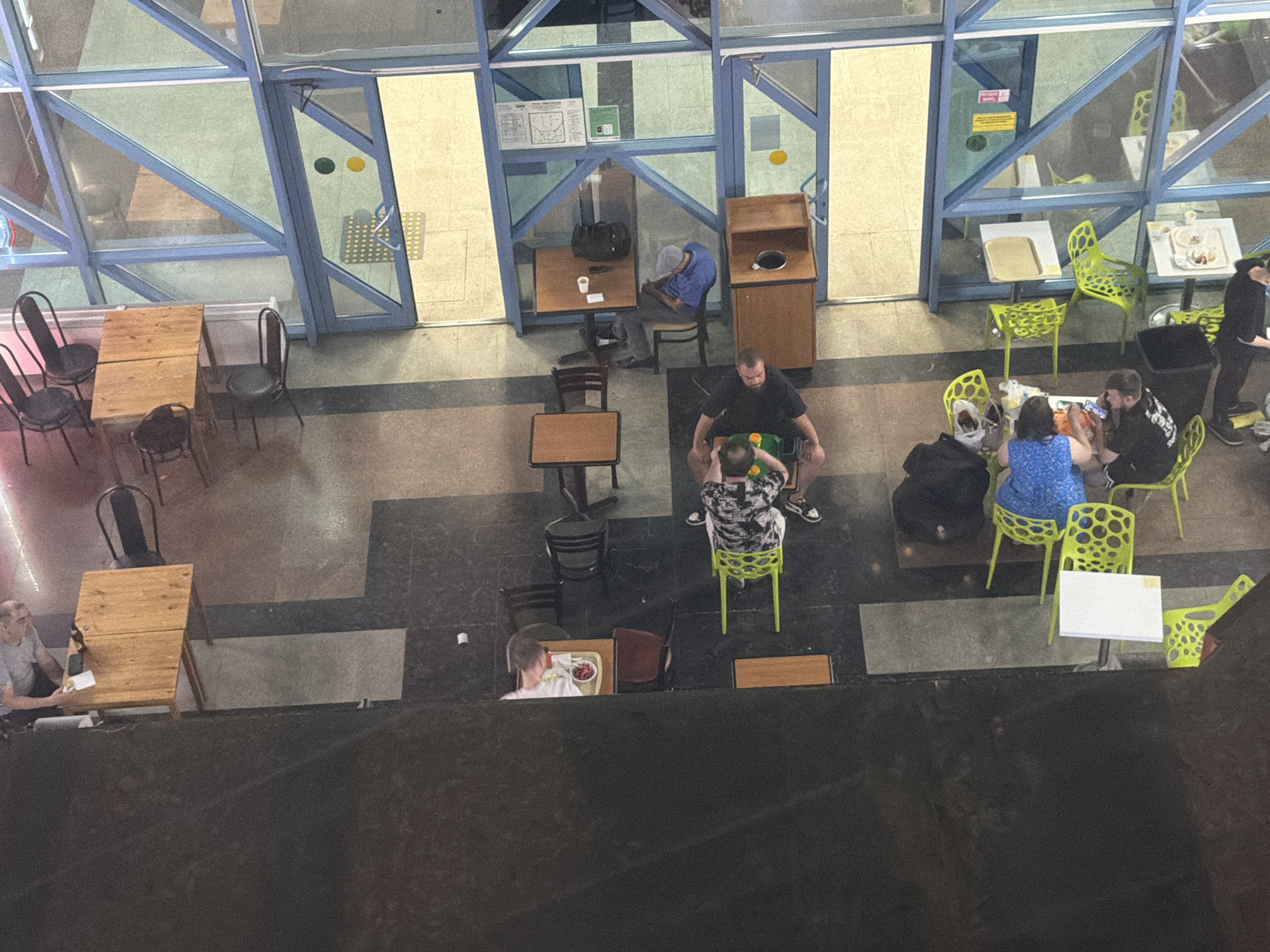 The image shows an overhead view of an indoor seating area, possibly a cafe or food court, located in Rostov-on-Don, Russia. The environment features a combination of tiled and dark flooring, with large windows and blue-colored structural elements dominating the background. Several tables and chairs are arranged throughout the space.
In the foreground, a man in a grey t-shirt is seated at a wooden table, holding a small piece of food. To his right, a brown chair is empty. Further back, two people, a man in a black t-shirt and a woman in a blue top, are seated at a table with a green chair. The man is holding a smartphone. Two green chairs are nearby, one occupied by a person wearing a patterned shirt and black pants.
In the middle ground, a person wearing a grey cap and blue shirt is seated at a table with a white cup. A black bag is on the floor next to the table. Another table with chairs is visible to the right, with plates of food on it. A person in a black t-shirt and pants is standing nearby.
In the background, there are more tables and green chairs. A wooden counter with a trash can is also present. The overall scene depicts a casual gathering or dining experience within an urban setting in Rostov-on-Don.