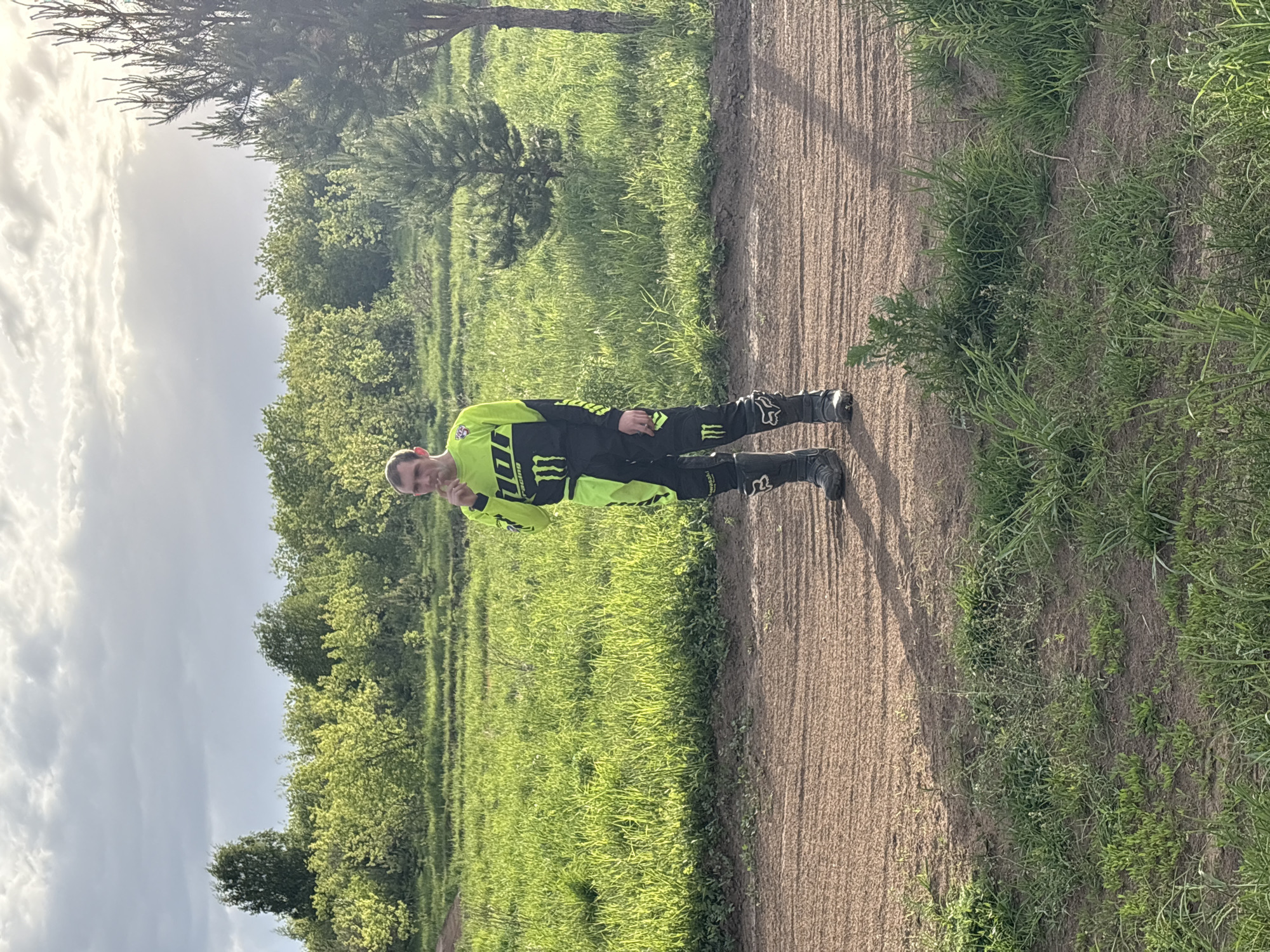 A person in motocross gear stands on a dirt path in a grassy area. The person is wearing a neon yellow and black racing suit with Monster Energy and Fox logos. The path is light brown dirt. The background consists of green grass and trees under a cloudy sky. The image's origin as Votkinsk, Russia is not verifiable.