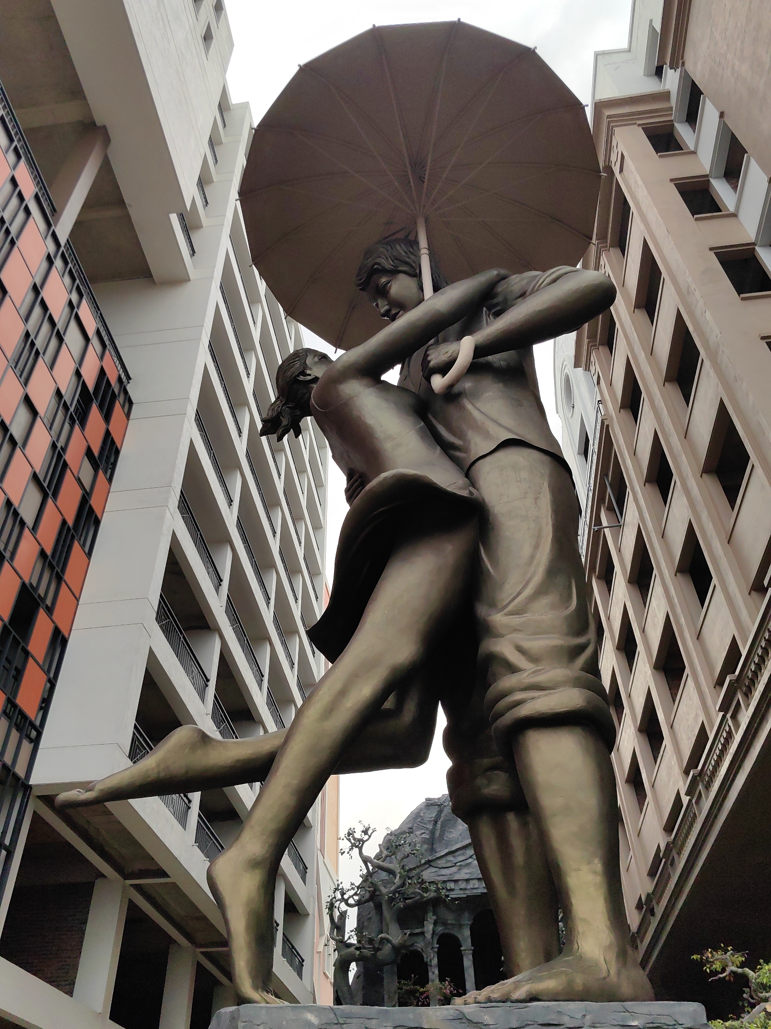 A low-angle shot depicts a bronze statue of a couple embracing under an umbrella, set against a backdrop of modern buildings in Tuan Chau Ward, Vietnam. The statue features a man holding an umbrella over a woman who is lifted off the ground in a romantic embrace. Both figures are barefoot and dressed in casual clothing. The surrounding architecture includes buildings with contrasting facades: one with a grid pattern of orange and black squares, another predominantly white with rows of balconies, and a third in a muted brown tone with window frames forming a rectangular pattern. Other sculptural elements are visible in the background near the base of the statue, adding depth to the scene. The overcast sky provides a neutral light.