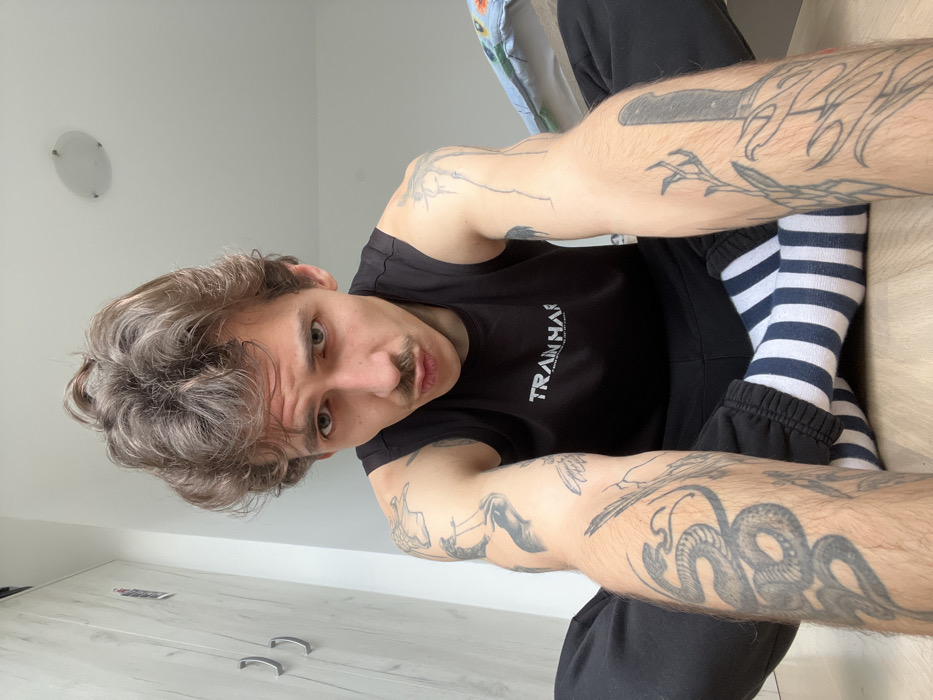 The image displays a male individual seated on a light-colored wooden floor indoors, facing the camera directly. He is a young adult with light brown, wavy hair, a mustache, and fair skin. He wears a black sleeveless tank top with "TRAIN HARD. LIVE FREE" in white text, black sweatpants, and white socks with thick black horizontal stripes. Numerous tattoos are visible on his arms and legs, including a large snake design on his right forearm (viewer's left), abstract patterns on his left forearm, and a design resembling a knife or dagger with organic elements on his left shin (viewer's right). He is in a cross-legged seated position with his arms extended towards the camera, indicating a self-portrait. His lips are pursed, and he maintains direct eye contact. The background consists of a light-colored wardrobe with silver handles on the left, a plain white ceiling with a circular flush-mounted light fixture in the upper right, and a multicolored patterned fabric, possibly a bedsheet, on the right side. The specific location in Rafailovići, Montenegro, cannot be determined from the visual evidence.