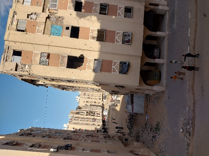 The image presents a daytime, outdoor scene dominated by several heavily damaged multi-story buildings, suggesting an urban environment that has experienced significant destruction. The overall impression is one of aftermath or ongoing hardship.
In the foreground and occupying the right side of the frame, a large, light-colored (beige or tan) multi-story residential building stands prominently. Its facade is severely compromised, with numerous large holes and missing sections revealing dark, empty interiors. Many windows are shattered, missing, or boarded up. Some windows retain their decorative white, patterned frames, contrasting with the raw damage. On one of the upper floors, a bright blue tarp or piece of fabric is visible, covering a portion of a window opening. The ground level of this building features arched openings, revealing what appear to be covered parking spaces, with at least one dark-colored car partially visible within one of these recesses.
Below this main building, a paved street runs horizontally across the lower part of the frame. The street surface appears worn and is bordered by areas covered in rubble, dirt, and debris. On this street, four individuals are visible. Closest to the viewer, one person in dark clothing stands with an arm extended towards another individual in lighter attire. Nearby, two smaller figures, likely children, are also standing on the street, one wearing a yellow/orange top and the other a darker top. They appear to be observing their surroundings or interacting with each other in a casual manner, amidst the scene of destruction.
Across the street, further into the mid-ground, another similar multi-story building is visible, also exhibiting extensive damage. Between this building and the foreground street, there is an open, unpaved area strewn with more debris and rubble. A white, tent-like structure is set up in this area, potentially serving as a temporary shelter or aid station. Beyond these immediate structures, more residential buildings, also appearing damaged, extend into the background under a clear, bright blue sky. The lighting suggests it is daytime, with visible shadows indicating the sun is relatively high. No discernible text is visible within the image.