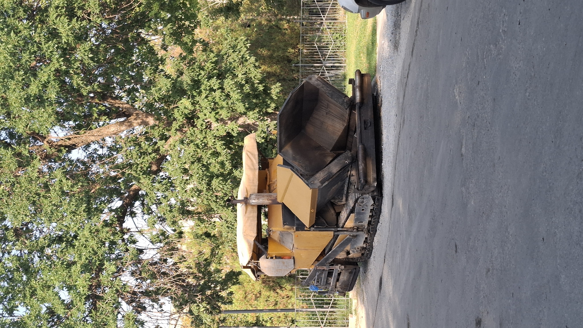 An asphalt paving machine, yellow and black, is parked on a paved surface. The machine has tracks and a large hopper at the rear, which appears to be empty. A beige canopy covers the top of the machine. In the background, there is a metal fence, green grass, and lush green trees. The scene appears to be an outdoor area within the city of Shymkent, Kazakhstan, possibly a street or construction site under renovation. The lighting suggests it is daytime. There are no people visible in the image.