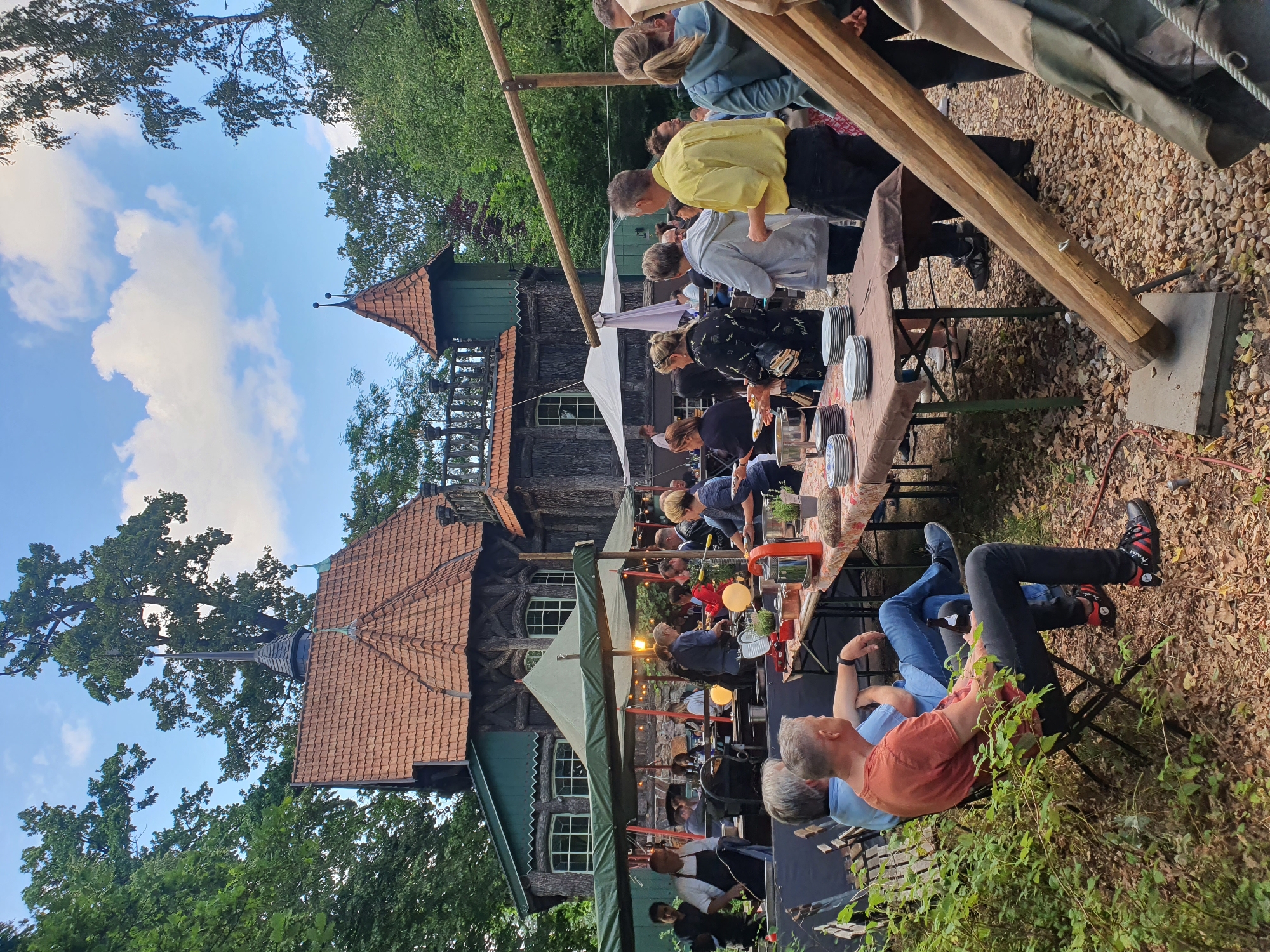 The image depicts an outdoor gathering or event taking place in a park-like setting with a rustic, fairytale-like building in the background. The building appears to be made of wood and features red tiled roofs and pointed towers, suggesting it might be a historical or themed structure. Several people are present, mingling, eating, and serving food around tables set up outdoors. Tents or awnings provide shade. The environment is green with trees and foliage. The presence of a distinctive building and the general ambiance suggests this location is likely a public space or a venue within Falkensee, Germany, possibly a restaurant, event space, or historical site.