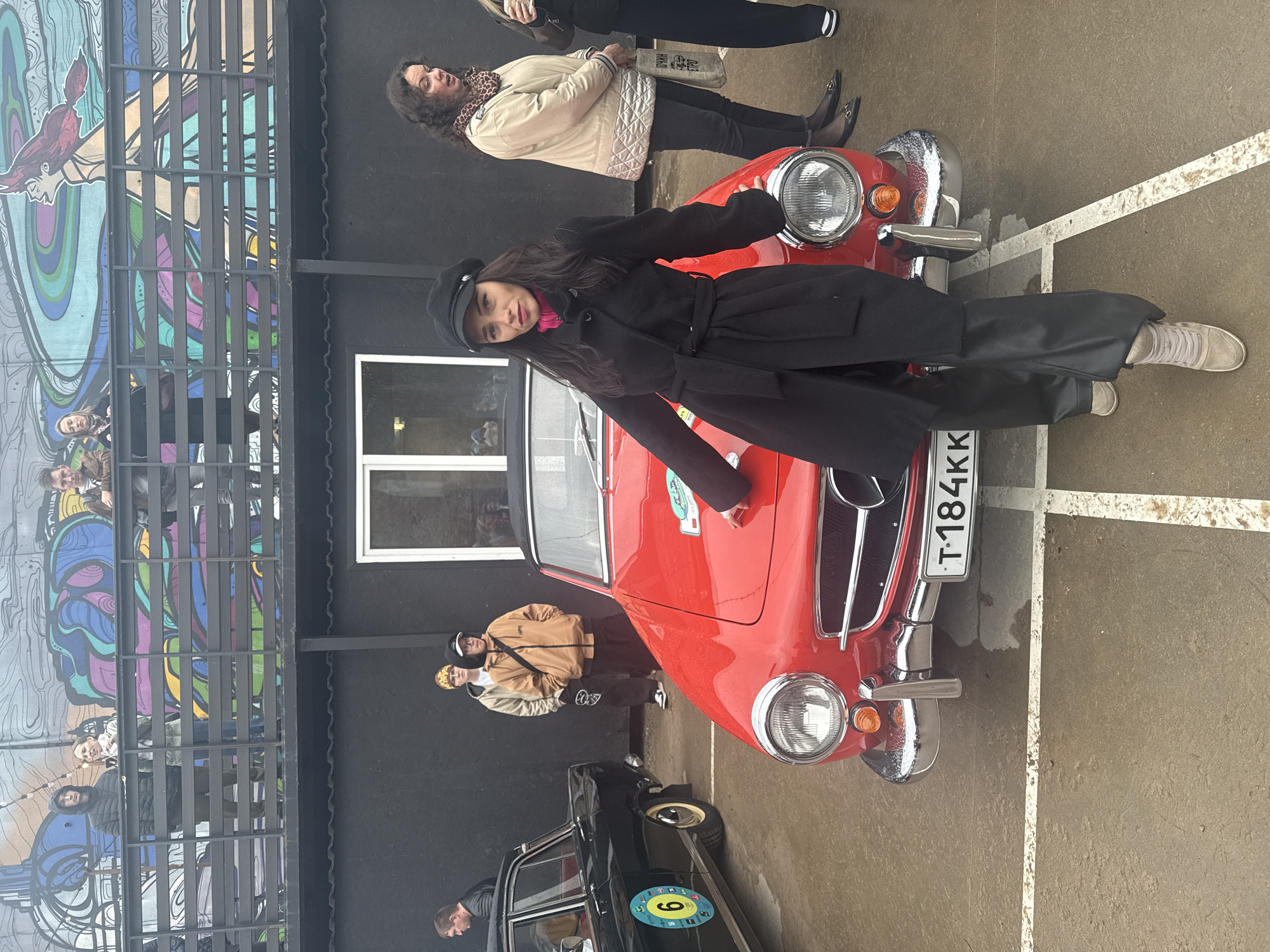 The image shows a red classic car, possibly a Mercedes-Benz, parked in front of a building with a distinctive mural in what appears to be a public space. A woman in a black coat and hat is posing on the car. A group of people are standing around the car, some looking at the camera. A black car is parked beside the red one. The environment suggests an urban setting, possibly in Saint Petersburg, Russia given the Russian license plate "T 184KK". The building has a black wall and a window, adjacent to an artistic mural depicting various figures and colors. The ground is paved and marked with white lines, indicating parking spaces.