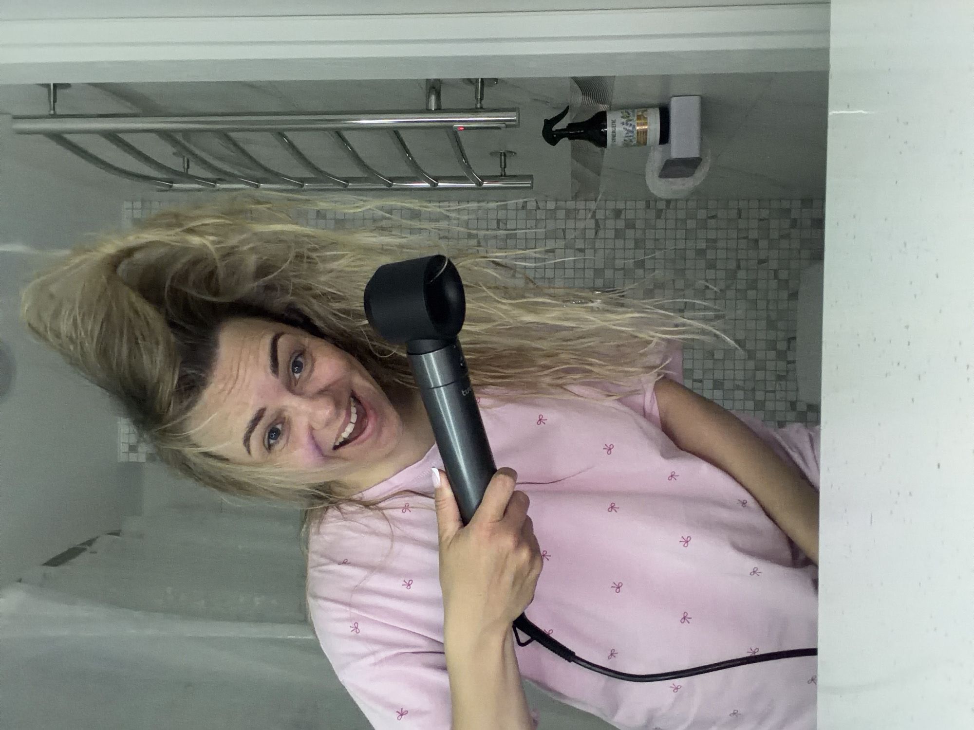 A woman is holding a Dyson Supersonic hair dryer in her right hand. She is smiling and facing the camera. She has blonde hair and is wearing a pink pajama top with a black trim and a white pattern. A black cord is attached to the bottom of the hair dryer. Behind her, there is a gray mosaic tile wall, a silver towel rack, and a spray bottle of "SYNERGETIC" cleaning fluid attached to the wall with a white dispenser. A white bathroom door is visible in the right part of the image, and a shower curtain or wall is on the left. The image is rotated to the left. There is no indication of the location being Votkinsk, Russia, in the image.