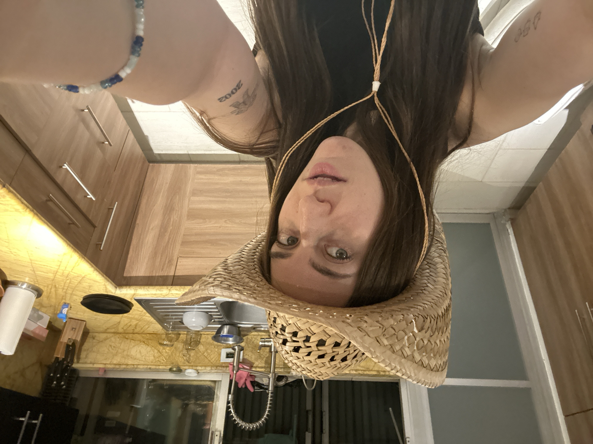 A selfie taken indoors shows a woman with long brown hair wearing a straw cowboy hat. She is upside down, positioned in front of a kitchen. The kitchen features wooden cabinets, a sink, a window, and various kitchen utensils. The woman has a tattoo on her arm and is wearing a beaded bracelet. The location is unspecified.