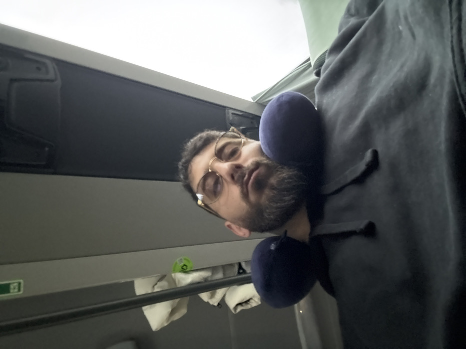An adult male is centrally depicted, seated within the interior of what appears to be a passenger transport vehicle. He possesses dark hair, a full beard, and wears gold-rimmed eyeglasses along with a dark-colored hooded sweatshirt. A navy blue U-shaped neck pillow is positioned around his neck. His gaze is directed towards the camera with a neutral to slightly narrowed expression. To his left, a light-colored, possibly padded object is visible hanging near a grey interior panel, which features a circular green sticker with indecipherable markings. The vehicle's ceiling is grey, with an overhead fixture present. To the right, an overcast sky is visible through a window, and a segment of a green upholstered seat or headrest is partially depicted. The male is seated; no other individuals or direct interactions are visible. There are no specific visual indicators within the image to confirm the location as Rosny-sur-Seine, France.