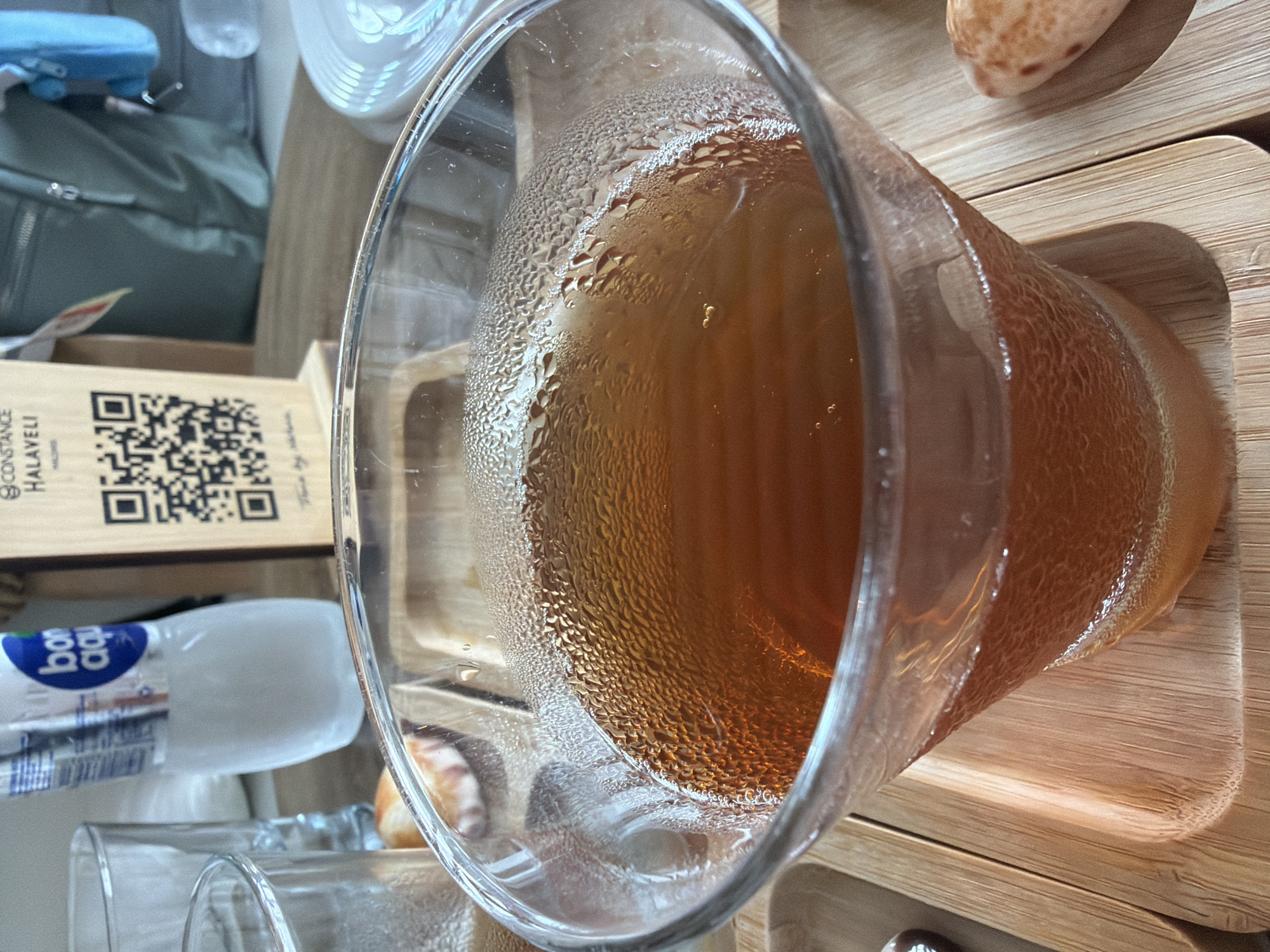 A close-up, eye-level shot showcases a glass bowl filled with a warm, amber-colored beverage, possibly tea, set on a wooden tray. The glass has a condensation layer on its outer surface, indicating the drink is hot. The beverage's surface is dotted with tiny bubbles, and a subtle sheen reflects the ambient light.
In the background, out of focus, is a wooden stand with a QR code and text that reads "CONSTANCE HALAVELI". To the left of the stand, a partially visible bottle of Bon Aqua mineral water stands upright. Adjacent to the bottle, two empty drinking glasses are present, one taller than the other.
Further in the background, a green backpack with a blue pouch is visible. The overall setting suggests an indoor environment, possibly a cafe or restaurant, in the Maldives, specifically at Constance Halaveli, a resort located on North Ari Atoll, Maldives. The scene is static, with no visible people or active interactions. The lighting appears to be natural or soft artificial light.