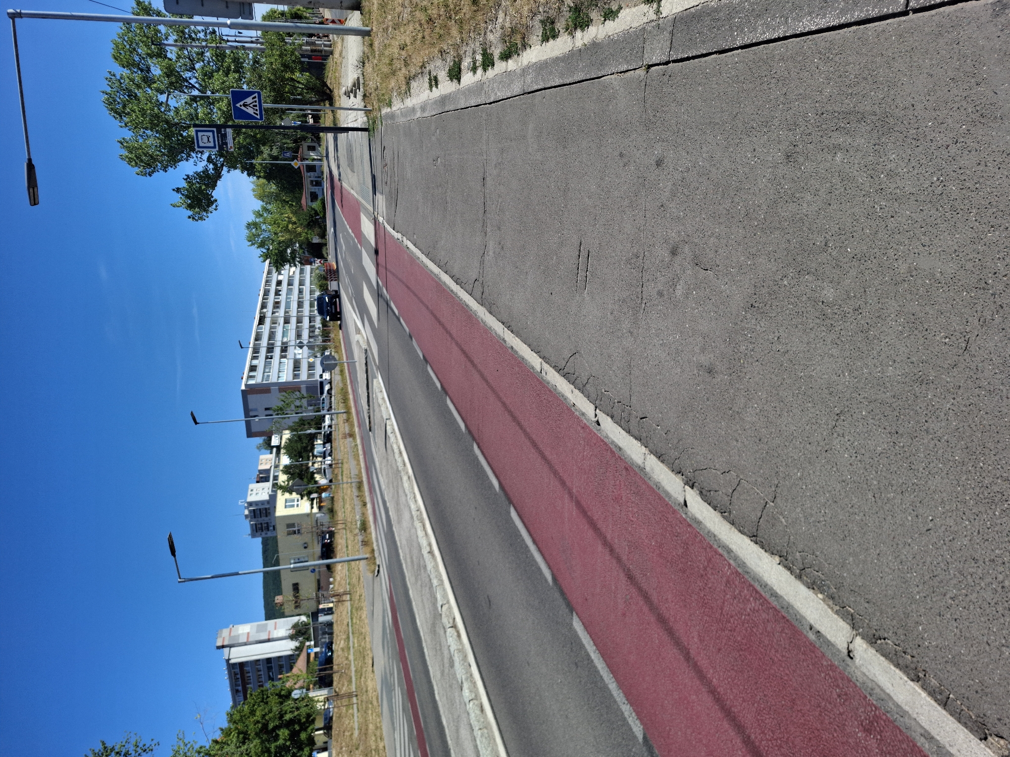 The image depicts a street scene in Bratislava, Slovakia, on a clear, sunny day. In the foreground, a cracked asphalt pedestrian path runs parallel to a street. The street itself is marked with a red-colored bike lane adjacent to the path and a grey lane for vehicular traffic. Across the street, a white pedestrian crossing is visible. Beyond the street, several multi-story apartment buildings and some smaller structures line the landscape, with trees and vegetation interspersed. A few cars are present on the street, and streetlights are positioned along the road. A pedestrian crossing sign is visible on the right side of the image. The overall impression is of an urban residential area.