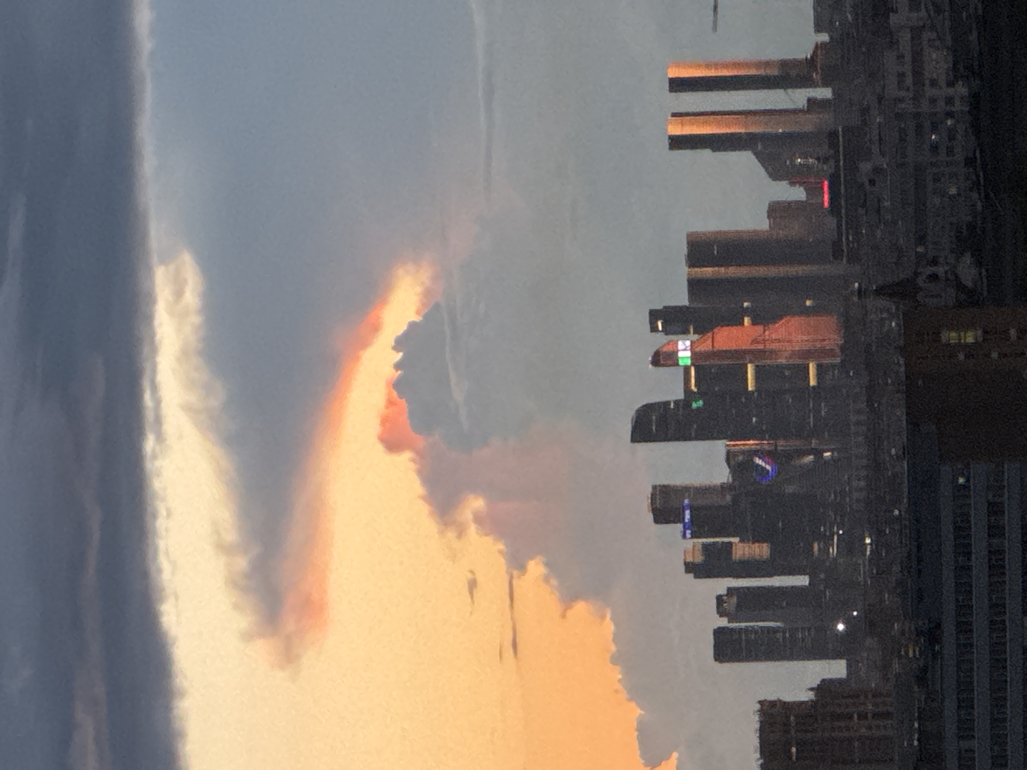 The image depicts a cityscape at dusk, likely Moscow, Russia, given the distinctive architecture of the skyscrapers. The sky is a dramatic blend of warm sunset hues and darker, storm-like clouds. The prominent skyscrapers, characteristic of Moscow City, are silhouetted against the sky. Some of the buildings are illuminated, with lights visible in windows and on facades. One skyscraper on the right is bathed in the warm, reddish-orange glow of the setting sun, reflecting off its glass surfaces. In the foreground, lower buildings and structures are visible, fading into darkness. The overall impression is one of urban grandeur juxtaposed with the powerful beauty of the natural elements.