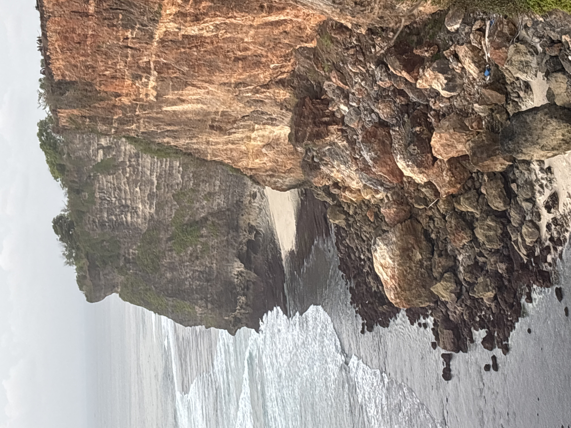 The image shows a dramatic cliff face overlooking the ocean in Kuta Selatan, Indonesia. The cliff is composed of layered rock, with patches of green vegetation clinging to its surfaces. At the base of the cliff, a rocky shoreline meets the incoming waves. The ocean is a muted gray-blue, with white foam from the waves washing ashore.
In the foreground, the cliff face is broken into a scree slope of large rocks and smaller stones. Some of these rocks are reddish-brown, likely due to iron content, while others are lighter in color. There is a small amount of debris visible amongst the rocks, including what appears to be blue plastic.
Further up the cliff, the rock layers are more visible, showing horizontal striations. Greenery is present on the ledges and in crevices. At the very top of the cliff, a group of people are visible, silhouetted against the sky, standing near the edge. Trees and bushes grow on the plateau at the top. The sky is overcast and the lighting suggests it is either early morning or late afternoon. The scene captures the rugged beauty of the natural landscape.