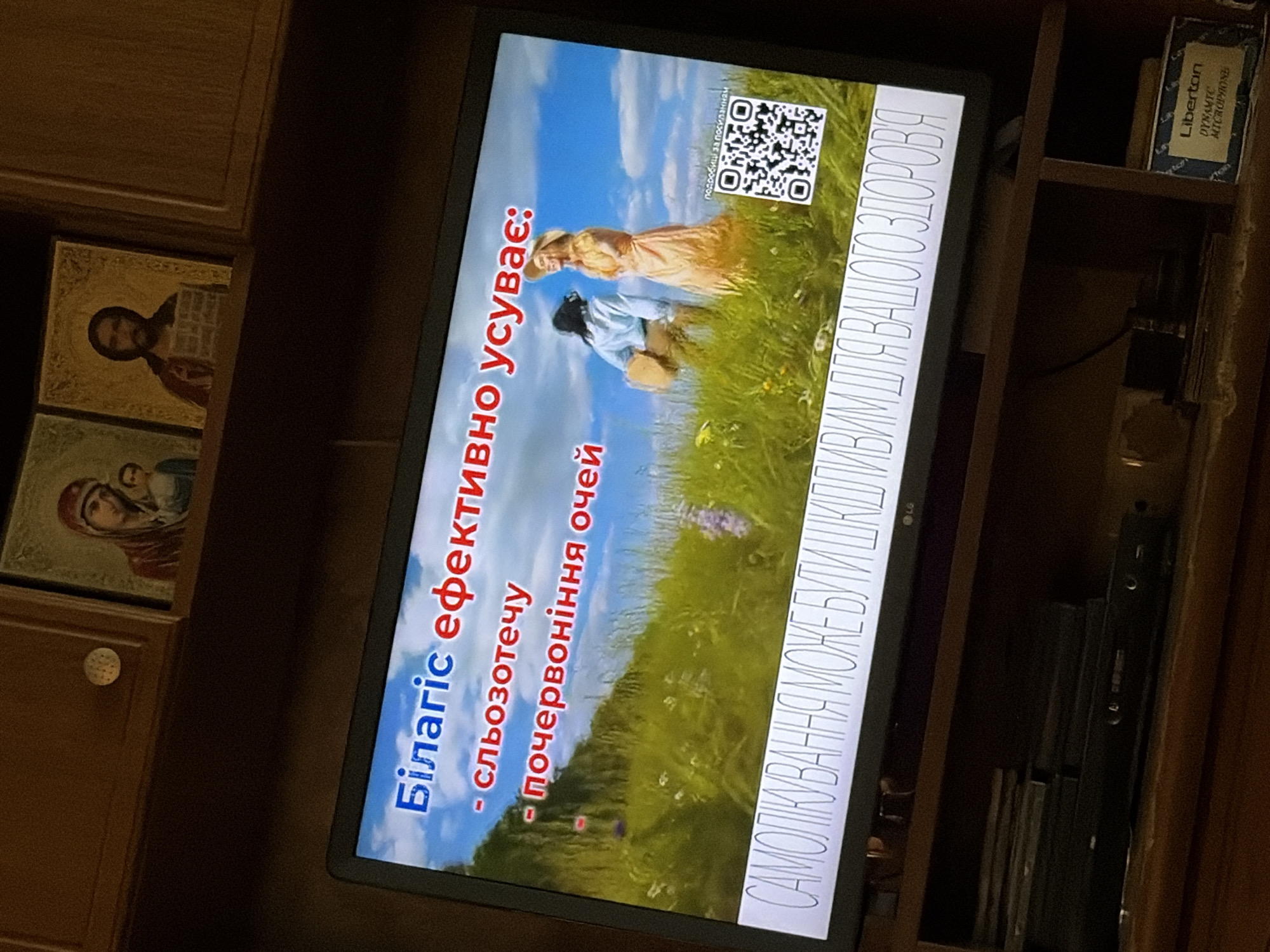 The image shows a television screen displaying a Ukrainian advertisement for "Bilagic". The text on the screen reads: "Білагіс ефективно усуває: - сльозотечу - почервоніння очей" which translates to "Bilagic effectively eliminates: - tearing - red eyes". Below this, a larger phrase reads "САМОЛІКУВАННЯ МОЖЕ БУТИ ШКІДЛИВИМ ДЛЯ ВАШОГО ЗДОРОВ'Я" which translates to "Self-medication can be harmful to your health". The advertisement features a pastoral scene with two people in a grassy field with a bright blue sky. A QR code is visible in the lower right corner of the screen. The television is placed on a wooden shelf. Above the television, two religious icons are visible. The overall environment appears to be a domestic setting, possibly a living room or a display shelf in a shop. The image is taken from a slight angle.