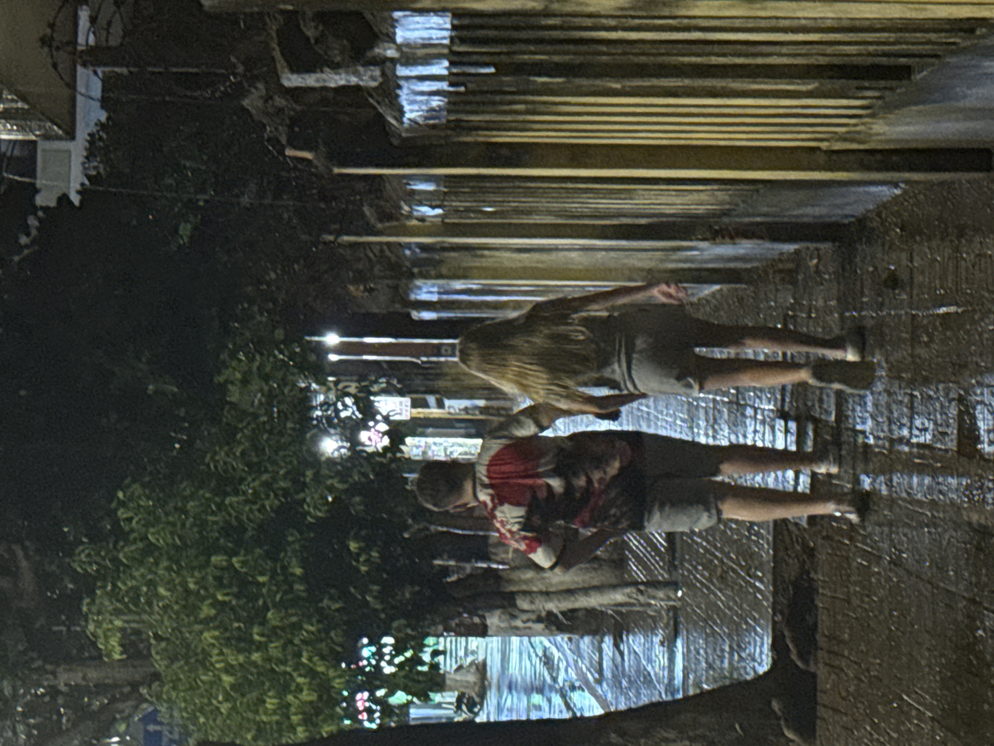 Two people walk on a dark, wet street in Bac Nha Trang Ward, Vietnam. The street is lined with trees and buildings. One person has long blonde hair and wears a gray shirt and shorts. The other wears a red shirt and dark shorts.