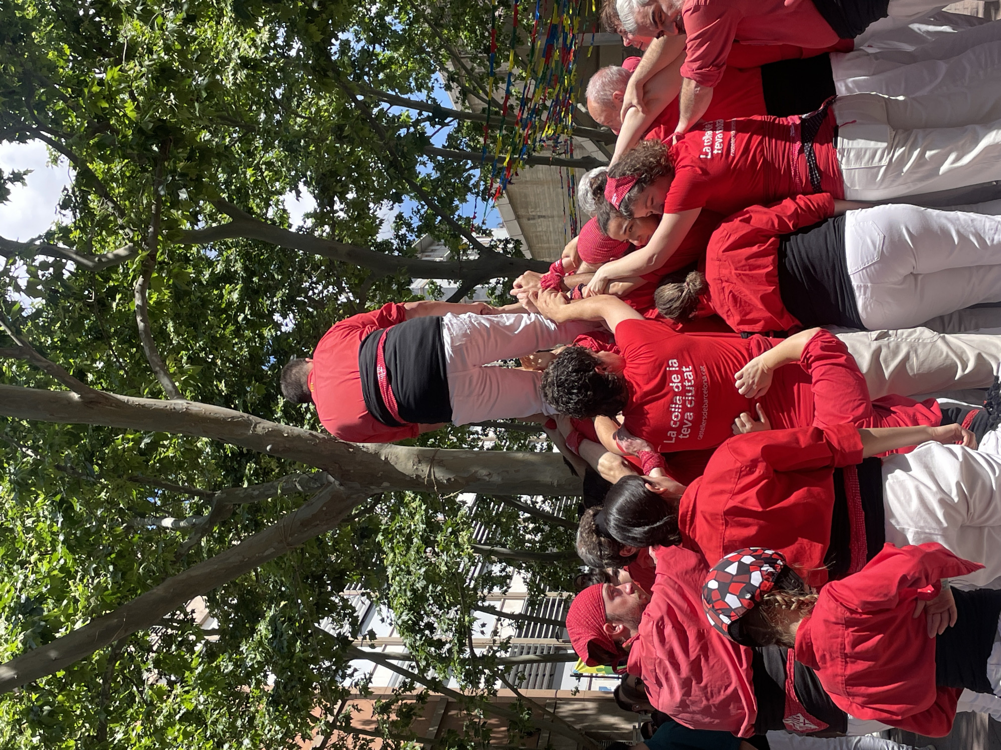 A medium shot depicts a human tower being built in Barcelona, Spain. The people forming the tower are wearing red shirts with "La colla de la teva ciutat castellersdebarcelona.cat" printed on them. They are also wearing white pants with black sashes. There are trees in the background and buildings visible in the distance. The scene seems to be happening outdoors during the daytime.
