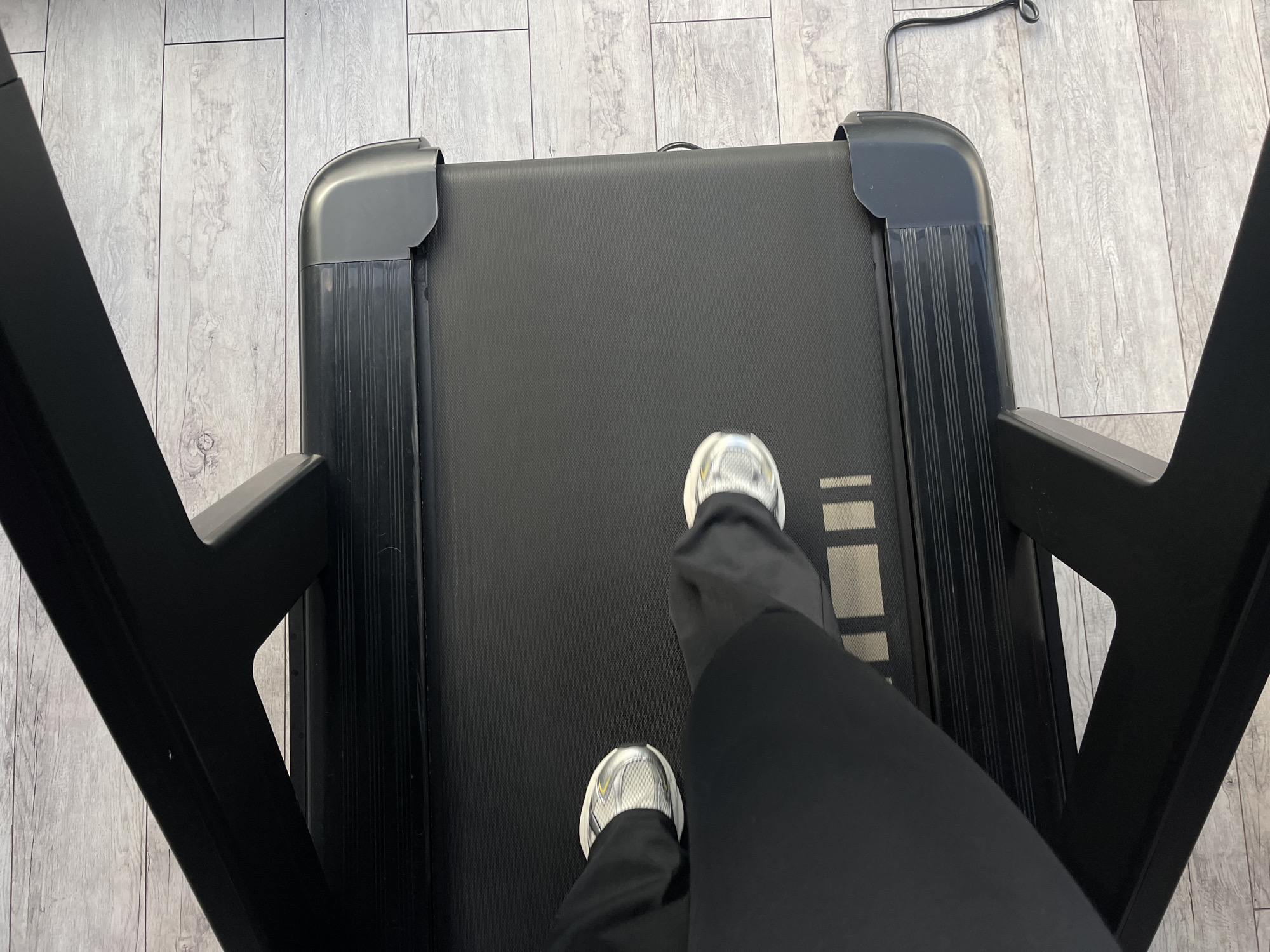 This is a top-down, eye-level shot of a person's legs and feet on a black treadmill. The person is wearing black pants and white athletic shoes. The treadmill is on a light gray, wood-plank style floor. The treadmill is black with gray end caps and has a textured surface on the side rails. A black power cord is visible trailing from the right side of the treadmill. The image does not contain any information that allows for its localization to Minsk, Belarus.