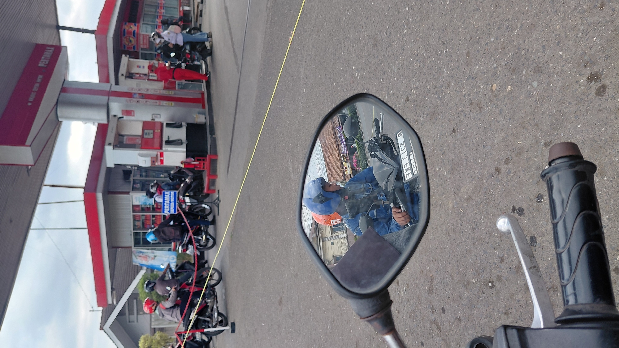 The image depicts a scene at a Pertamax gas station in Rejasari, Indonesia. In the foreground, a motorcycle's handlebar and a portion of its side mirror are visible. The side mirror reflects a person wearing a blue helmet and a denim jacket, sitting on a motorcycle. Behind the motorcycle, several other motorcycles are parked, and some individuals are seen interacting with gas pumps. A red and white Pertamax sign is prominently displayed on the gas station structure. The environment appears to be an outdoor area with paved ground, under an overcast sky. The overall impression is one of everyday activity at a local gas station.