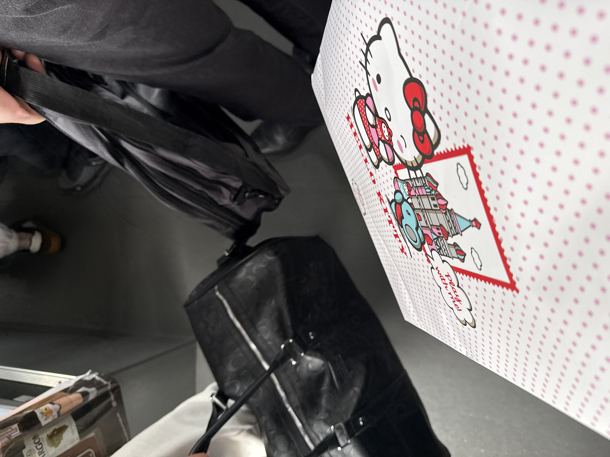 A high-angle shot depicts a person carrying a black duffel bag alongside a white shopping bag adorned with Hello Kitty and a castle illustration. The bag has a pink dot pattern. Other items include a black shoulder bag and a box with visible printed text. The scene is indoors, likely on a train or bus, as suggested by the flooring and nearby seats. It's impossible to determine the specific location in Khimki, Russia, or any interactions without more context.