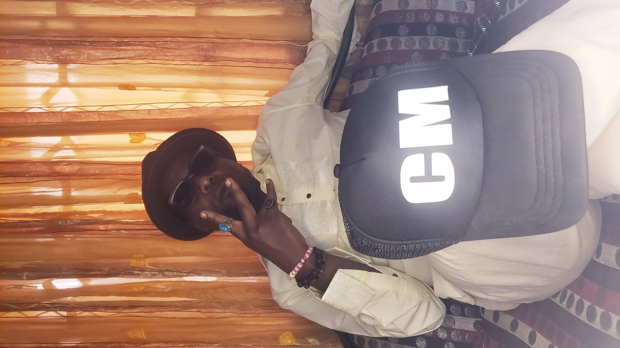 An African man, wearing a brown hat and sunglasses, sits against a backdrop of orange curtains. He wears a light-colored shirt and bracelets on his wrist, and multiple rings on his fingers. He holds two fingers up in a peace sign gesture. Beside him sits a black baseball cap with white lettering that reads "CM". The background also includes patterned pillows. The location is Narayi, Nigeria.