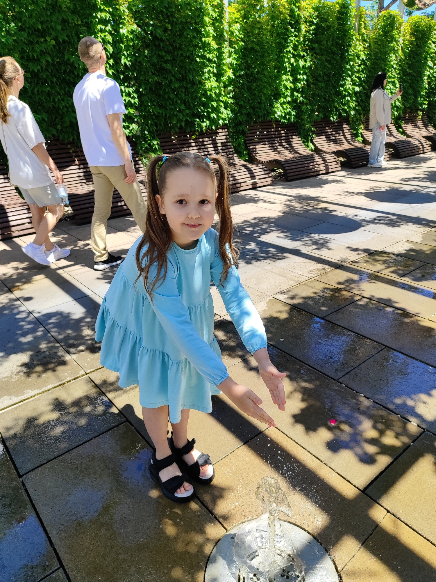 A vibrant outdoor scene captures a young girl playing in a public space on a sunny day. In the foreground, a girl with brown pigtails, dressed in a light blue tiered dress and black sandals, smiles towards the camera. Her hands are cupped over a small, ground-level fountain from which water is actively spraying, wetting the surrounding light-colored paving stones.
In the background, several blurred figures can be seen. To the left, a woman in a white t-shirt and shorts carries a water bottle while walking alongside a man in a white t-shirt and khaki pants. To the right, another woman in a light-colored jacket and jeans stands near a series of slatted wooden benches, holding a phone as if taking a picture.
The area behind the benches features a tall wall covered densely with lush green ivy, creating a natural green backdrop. Strong shadows from overhead trees or structures are cast across the paved ground, indicating bright sunshine, likely mid-day. The setting appears to be a well-maintained park or recreational area, consistent with the context of Krasnodar, Russia. The girl's expression suggests joy and engagement with the water. No visible text is present in the image.