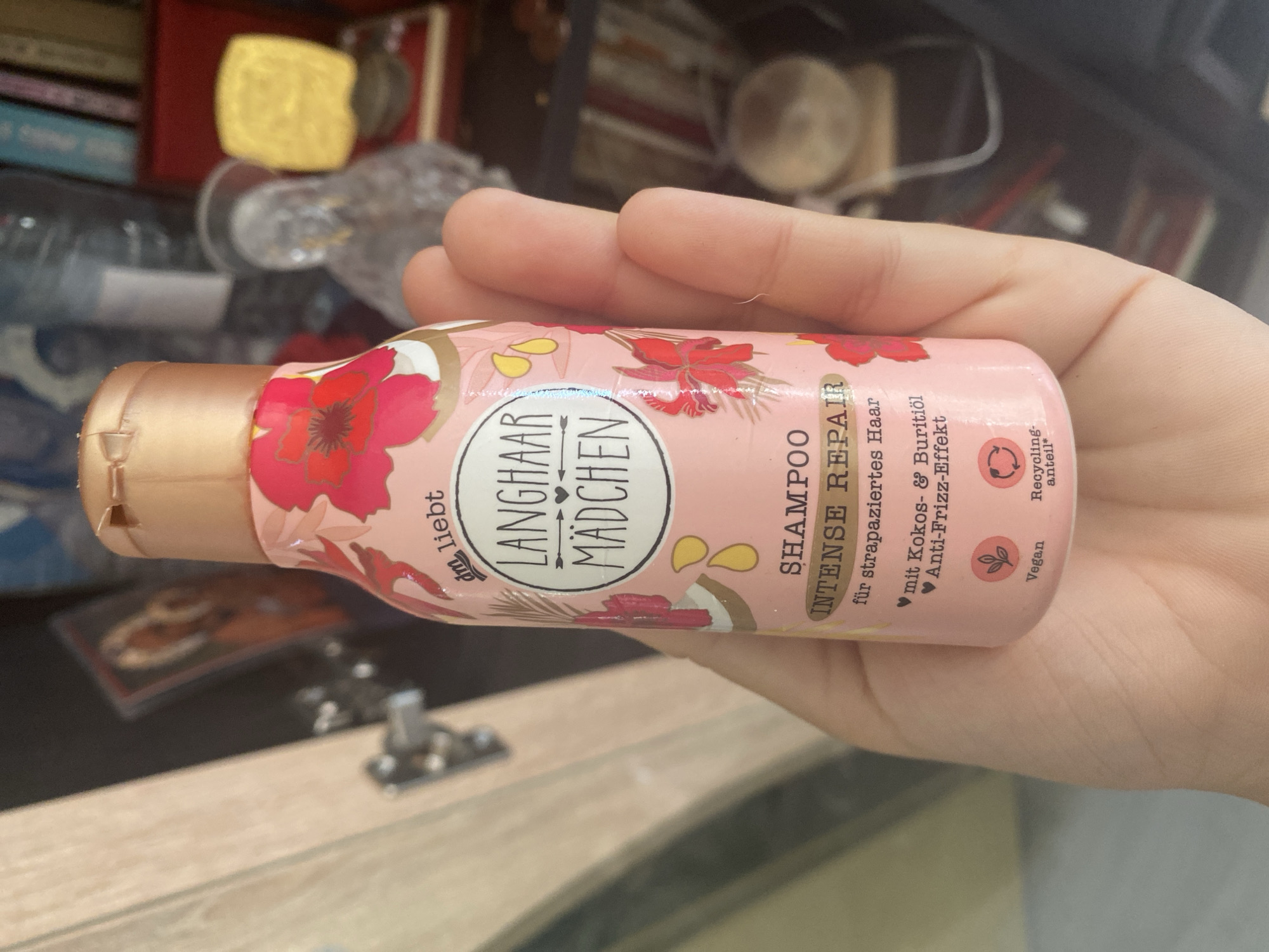 A person's hand is holding a pink bottle of "Langhaar Mädchen Intense Repair Shampoo" with a floral design. The bottle has a rose-gold cap and text that says "dm liebt LANGHAAR MÄDCHEN SHAMPOO INTENSE REPAIR für strapaziertes Haar mit Kokos- & Buritiöl Anti-Frizz-Effekt". Below this, there are icons indicating "Vegan" and "Recycling-Anteil". The background is out of focus and shows a bookshelf with various items, including what appears to be a religious icon, a clear glass figurine, and books. The overall setting suggests an indoor environment, possibly a home or a shop, but there is no information to determine its specific location in Blagoevgrad, Bulgaria.